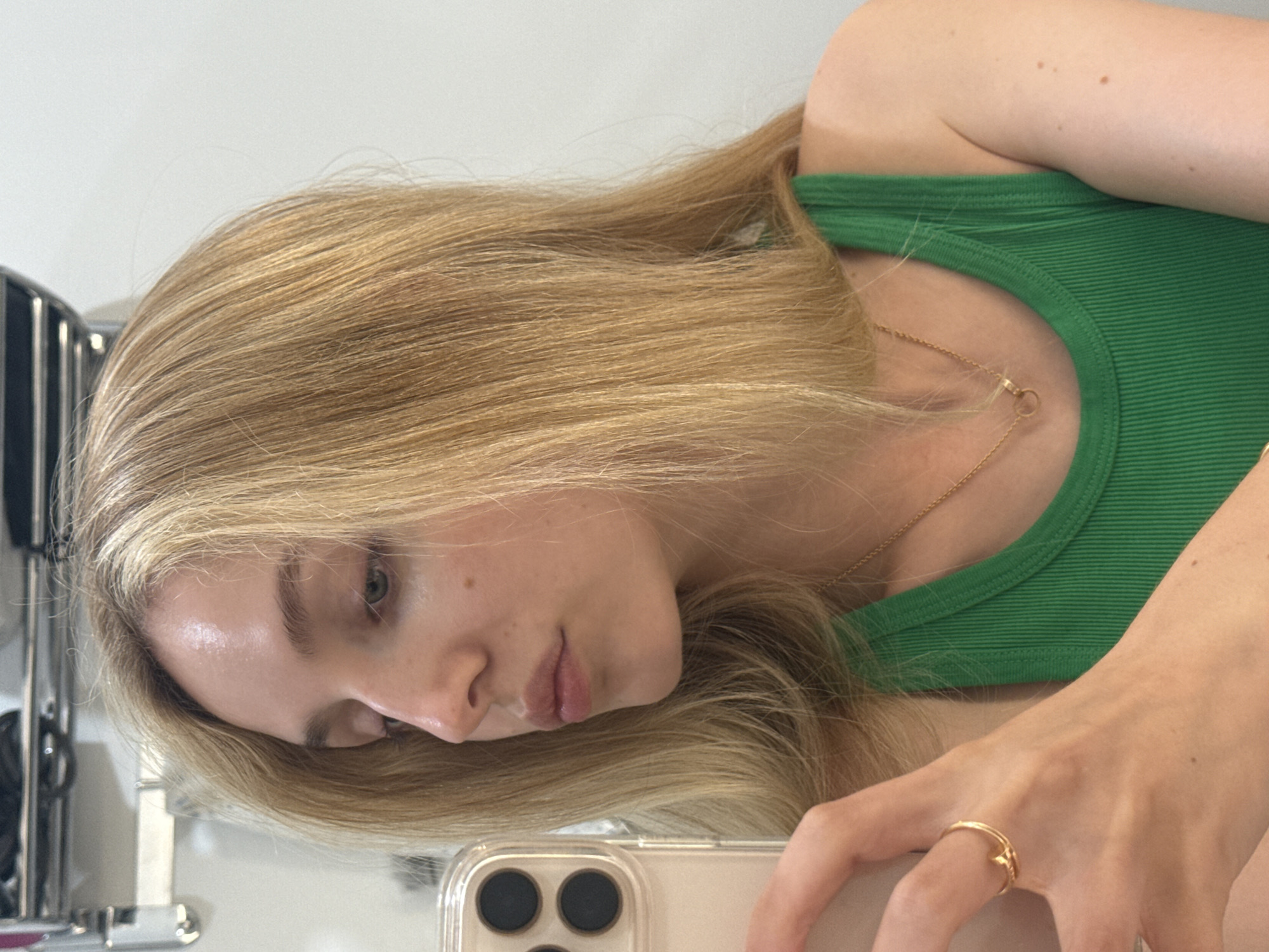 A young woman with shoulder-length blonde hair is taking a selfie with her phone in a bathroom. She is wearing a green tank top and a gold necklace. Her left hand is holding the phone, and she is wearing a gold ring on her index finger. The background is a white wall with some shelving above.