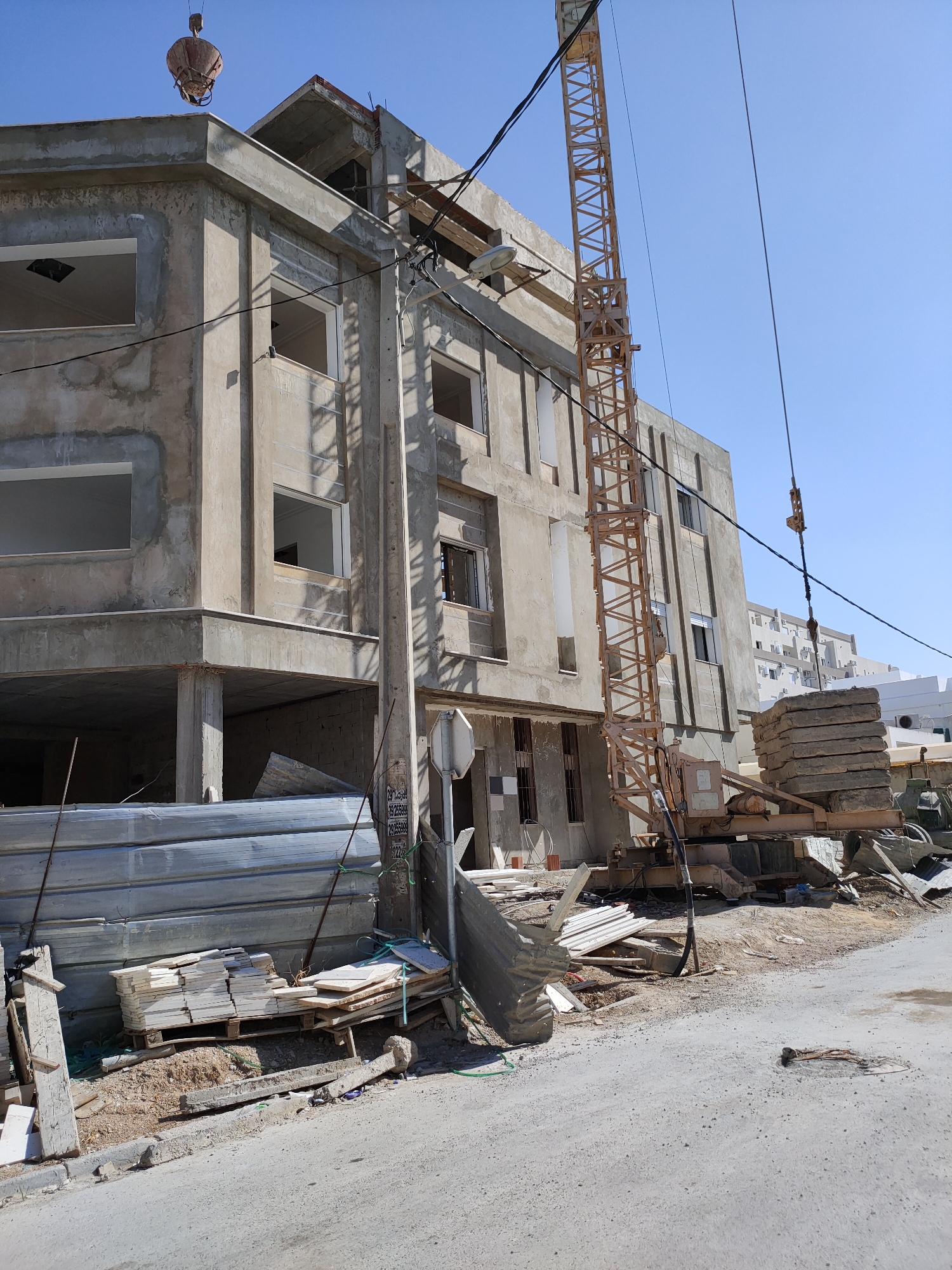 A construction site in Unknown, Tunisia, features a multi-story building under construction. The building is primarily concrete with unfinished facades and open window frames. A yellow tower crane is positioned next to the building, with its boom extending upwards and over. Several loads of construction materials, including stacked concrete slabs and piles of what appear to be tiles or flat stone pieces, are visible on the ground and near the crane. Metal fencing and other debris are scattered around the site. Power lines crisscross the scene, and the sky is clear and blue. In the foreground, a rough, unpaved road leads away from the construction site.