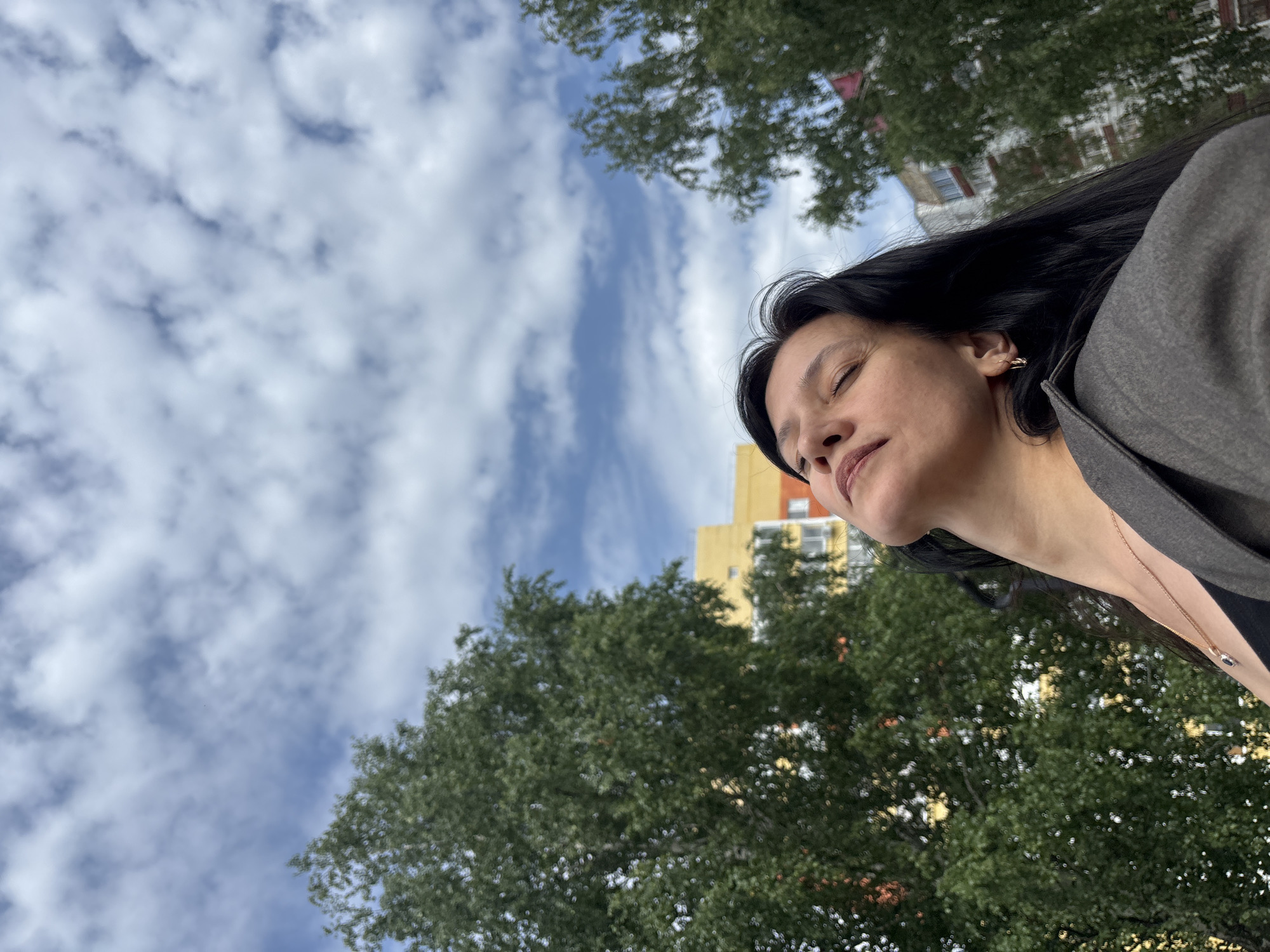 An overhead shot, from a low angle, shows a woman with dark hair and closed eyes, tilted back with her face towards the sky. She is wearing a gray blazer and a black top, with a delicate gold necklace. The background features a sky filled with white, puffy clouds, and some trees. In the distance, a multi-story residential building with yellow and orange accents is visible. The scene suggests an outdoor setting in the city of Chaikovsky, Russia, with the residential building being a characteristic element of Russian urban landscapes.