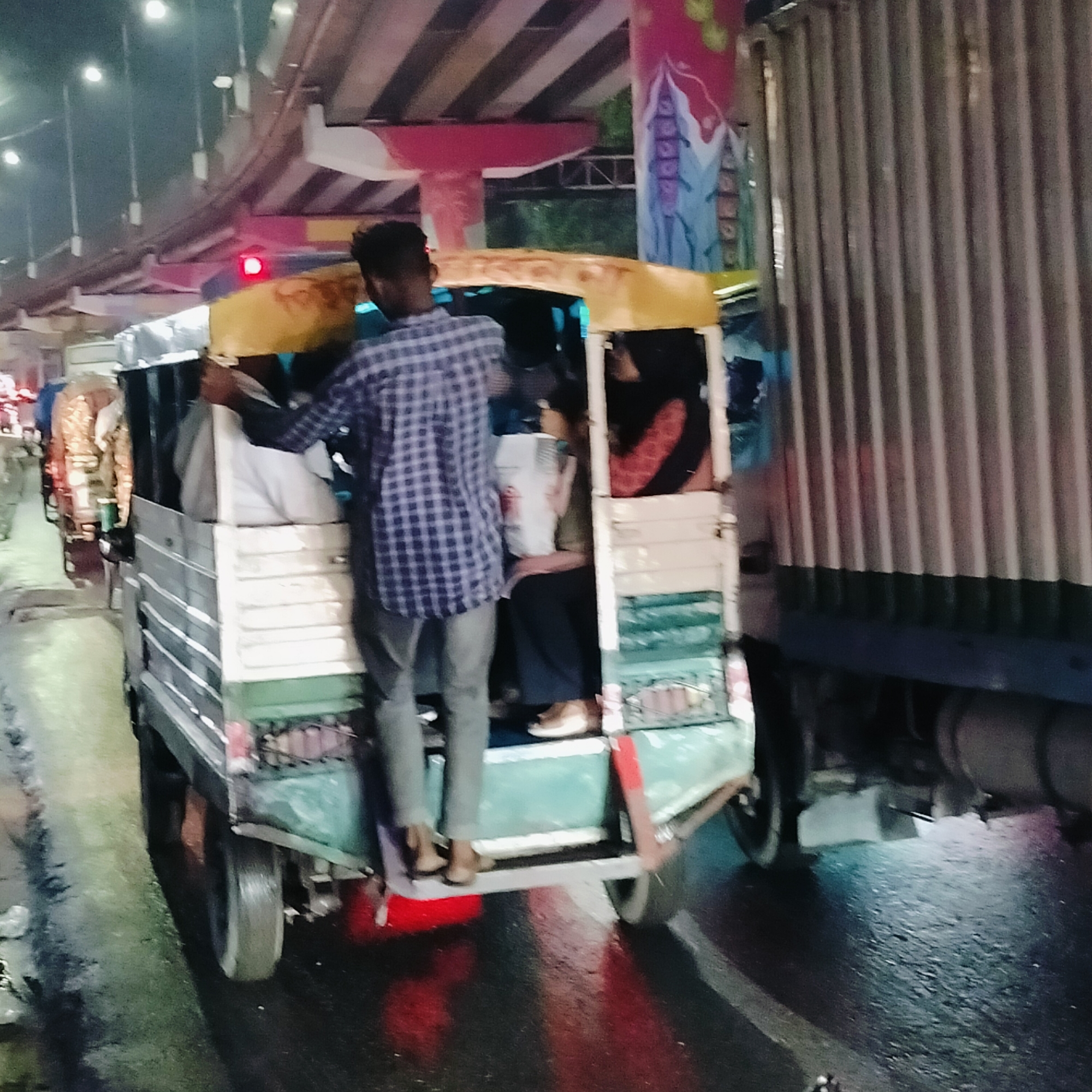 The image shows a crowded CNG auto-rickshaw in Dhaka, Bangladesh, at night. A man is standing on the footboard of the rickshaw, holding onto the side for support. Several passengers are inside. The scene is under an overpass with artistic painting on the support pillars. A large truck is to the right of the auto-rickshaw. The road is wet, reflecting the streetlights. The general atmosphere seems urban and busy.
