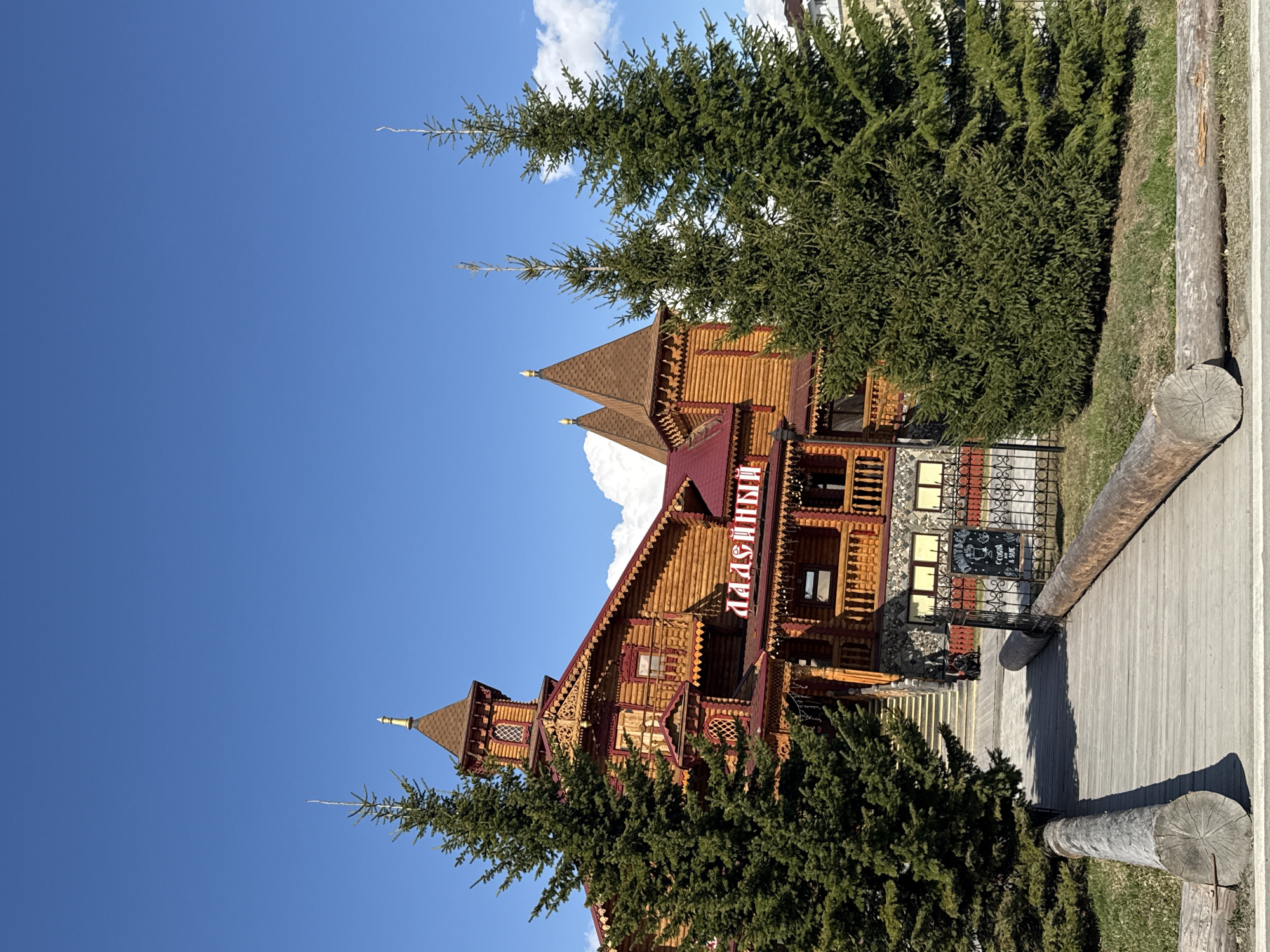 This image showcases a large, elaborately designed wooden building under a bright blue sky with scattered white clouds, characteristic of a sunny day. The structure, located in Tobolsk, Russia, is constructed from logs in a traditional Russian architectural style, featuring multiple stories, intricate carved detailing on its wooden facade, and prominent red and brown roofs culminating in pointed spires adorned with golden finials.
Several tall, dense evergreen trees, likely spruce or fir, are prominently featured in the foreground and midground, partially framing and obscuring parts of the building. A wooden boardwalk or pathway extends along the right side of the image, bordered by large, horizontally placed log barriers. Patches of sparse green grass are visible adjacent to the pathway.
The building's facade prominently displays the name "МАЛЬВИНА" (Malvina) in red Cyrillic letters. Near what appears to be an entrance, behind a decorative black metal fence, there is a smaller dark sign with some white, largely illegible text, hinting at advertised services or products, possibly including "COFFEE" or "COGNAC". No people are visible, and the scene presents a tranquil view of the architectural landmark amidst natural surroundings. The bright sunlight indicates daytime.