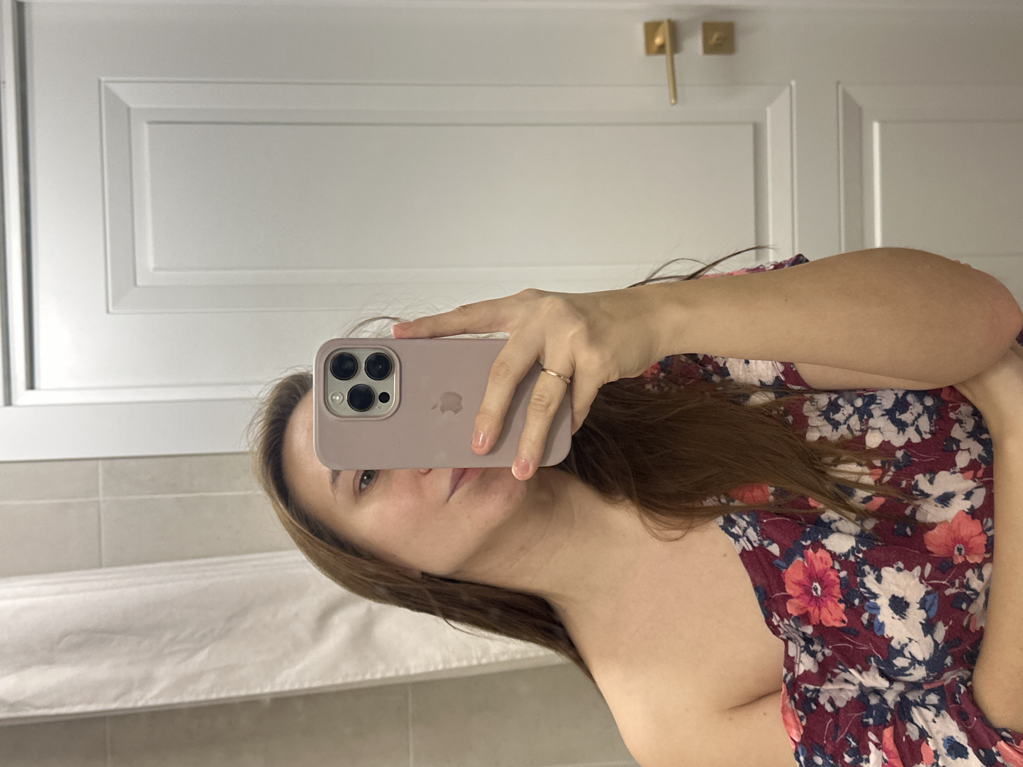 A woman holds a smartphone with a lilac case up to a mirror. She is taking a selfie. She has light brown hair and is wearing a floral dress. The reflection in the mirror shows white cabinets with gold handles and a light-colored tiled wall. The image was taken in an indoor location, likely a bathroom. There is no indication the image was taken in Kharnavy, Russia.