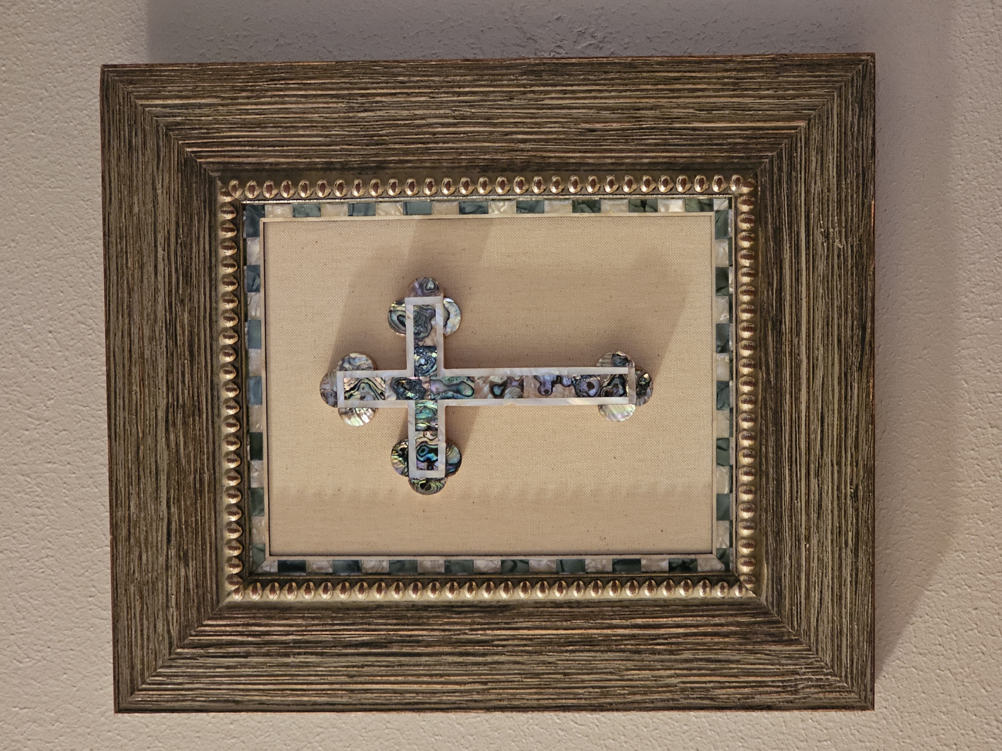 A framed abalone mosaic cross hangs on a textured beige wall. The cross is centered within a mat of pale beige fabric and is surrounded by decorative borders. The inner border consists of a strip of small, alternating square abalone tiles in shades of green and blue, followed by a border of small, gold-colored beads. The outer frame is made of wood with a distressed, textured finish, appearing in shades of brown and grey. The lighting casts a shadow on the wall below the bottom edge of the frame. The image does not depict any specific location in Amarillo, United States of America, nor does it contain any people or actions.