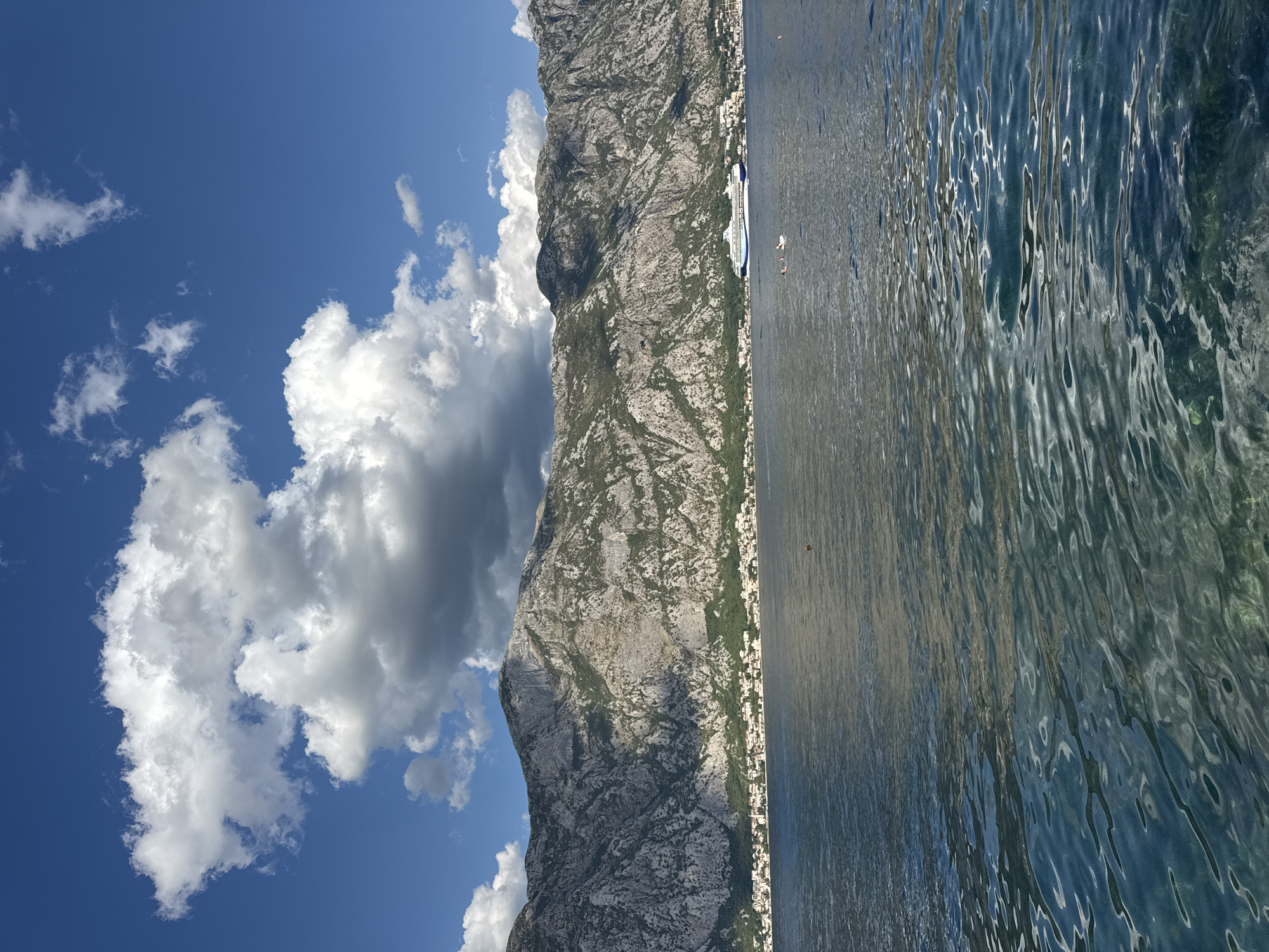 The image shows a body of water with a town nestled at the base of a rocky mountain range. The water in the foreground is clear and shows ripples, with glimpses of what appears to be the seabed or aquatic plants beneath the surface. Further out, the water is a deeper blue and calmer. In the middle ground, a cruise ship is visible docked or cruising near the coastline. A small town with white buildings, some topped with red roofs, lines the shore, with a church steeple being a prominent feature. The mountain range behind the town is rugged and rocky, with sparse vegetation. The sky above is a vibrant blue with scattered white and gray clouds. The scene is illuminated by bright sunlight. The location is identified as Prčanj, Montenegro.