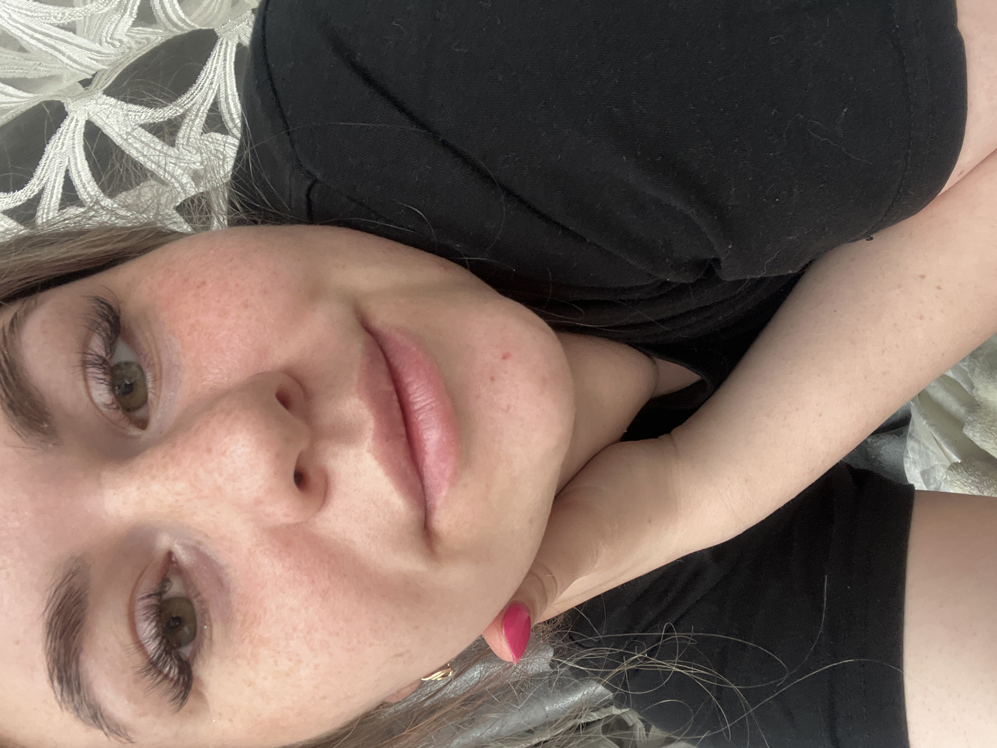 A close-up, high-angle portrait shot features a woman lying down, looking directly at the camera. She has light brown hair, which frames her face, and light brown or hazel eyes with prominent, dark eyelashes. Her eyebrows are well-defined. Numerous freckles are visible across her nose, cheeks, and forehead. Her lips are a soft pinkish-mauve color.
She is wearing a plain black t-shirt or top. Her left hand is resting under her chin or jawline, and her fingernails are painted a vibrant fuchsia pink. In the background, to her left, a pillow or blanket with an intricate white geometric pattern on a darker fabric is visible. To her right, a plain, lighter fabric is partially seen. The overall impression is one of casual relaxation, possibly indoors, with a neutral to slightly smiling expression. There is no visible text. The location context provided is Chaikovsky, Russia.