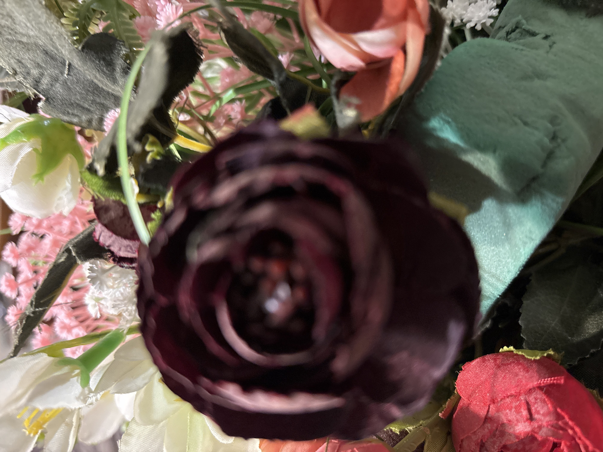 The image displays a close-up, top-down view of a dense arrangement of artificial flowers and foliage. A large, dark maroon or deep purple flower is prominent in the center, appearing significantly out of focus with blurred, spiraling petals.
Surrounding this central bloom are various other fabric and plastic flowers. In the upper right quadrant, a peach or light orange rose-like flower is visible, while in the bottom left, several white flowers with distinct yellow stamens are present. A bright red, textured fabric flower peeks into the frame from the bottom right. The background is filled with softer pink, clustered blossoms, and small, delicate white flowers resembling baby's breath, interspersed with various green leaves and stems. A textured, teal or light green fabric element can be seen towards the upper right, and a dark grey, somewhat fuzzy element is visible on the left side, possibly another artificial leaf or decorative component.
The image captures a static floral display, likely an indoor decoration. The lighting is soft and diffused, offering no specific cues about the time of day or outdoor weather conditions. The overall focus of the image is soft, with the central flower being particularly blurry. There are no people, activities, or visible text in the scene. The artificial nature of the flowers is evident from the fabric textures and construction.
