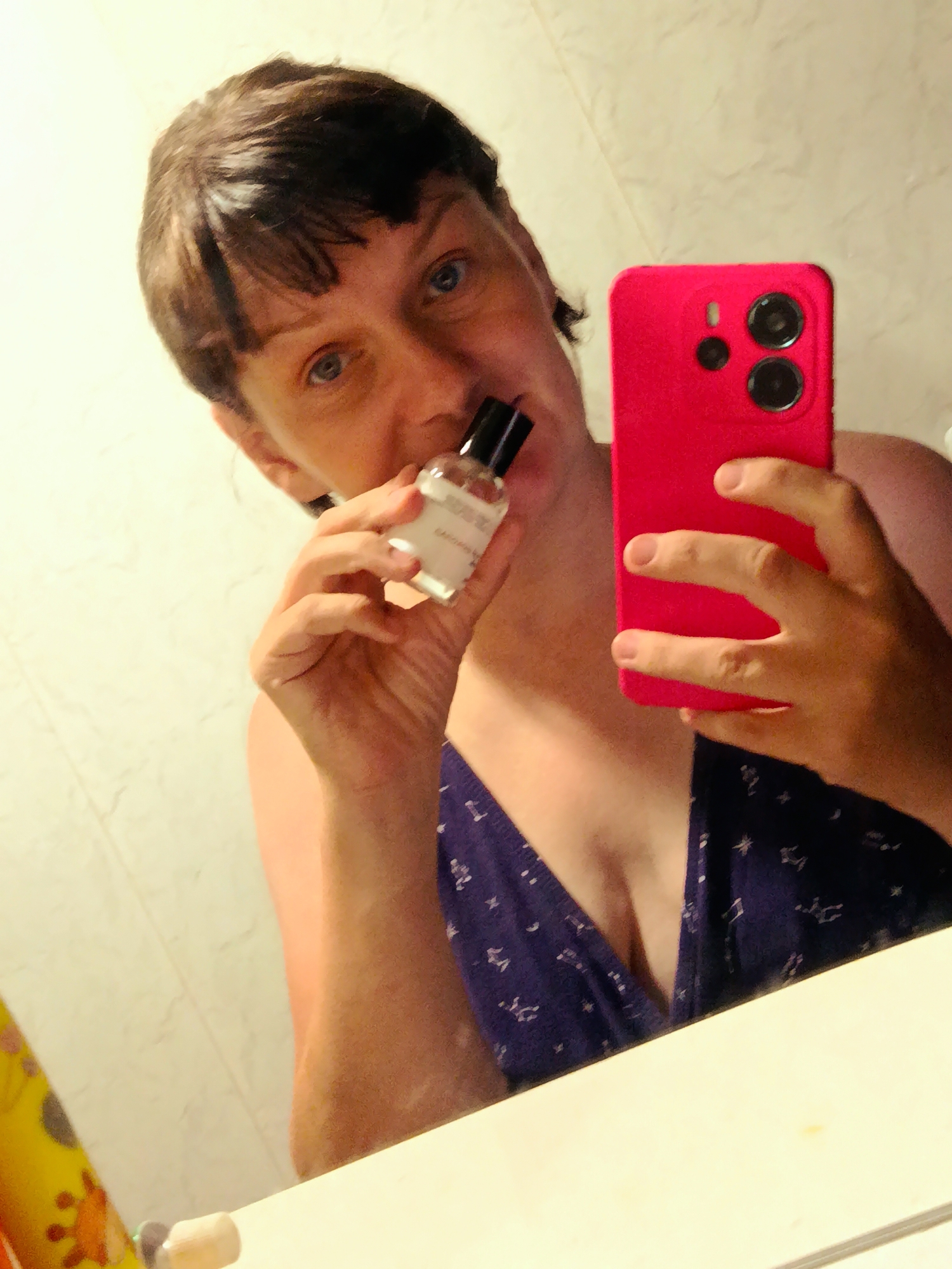 The image shows a person holding a small perfume bottle and a pink smartphone in a bathroom. The person has short, dark hair and blue eyes, and is wearing a dark blue dress with a star pattern. The bathroom has beige tiled walls and a mirror reflecting the scene. The person is in the process of applying perfume. The image does not provide any information about the location in the city of Фоки, Russia.