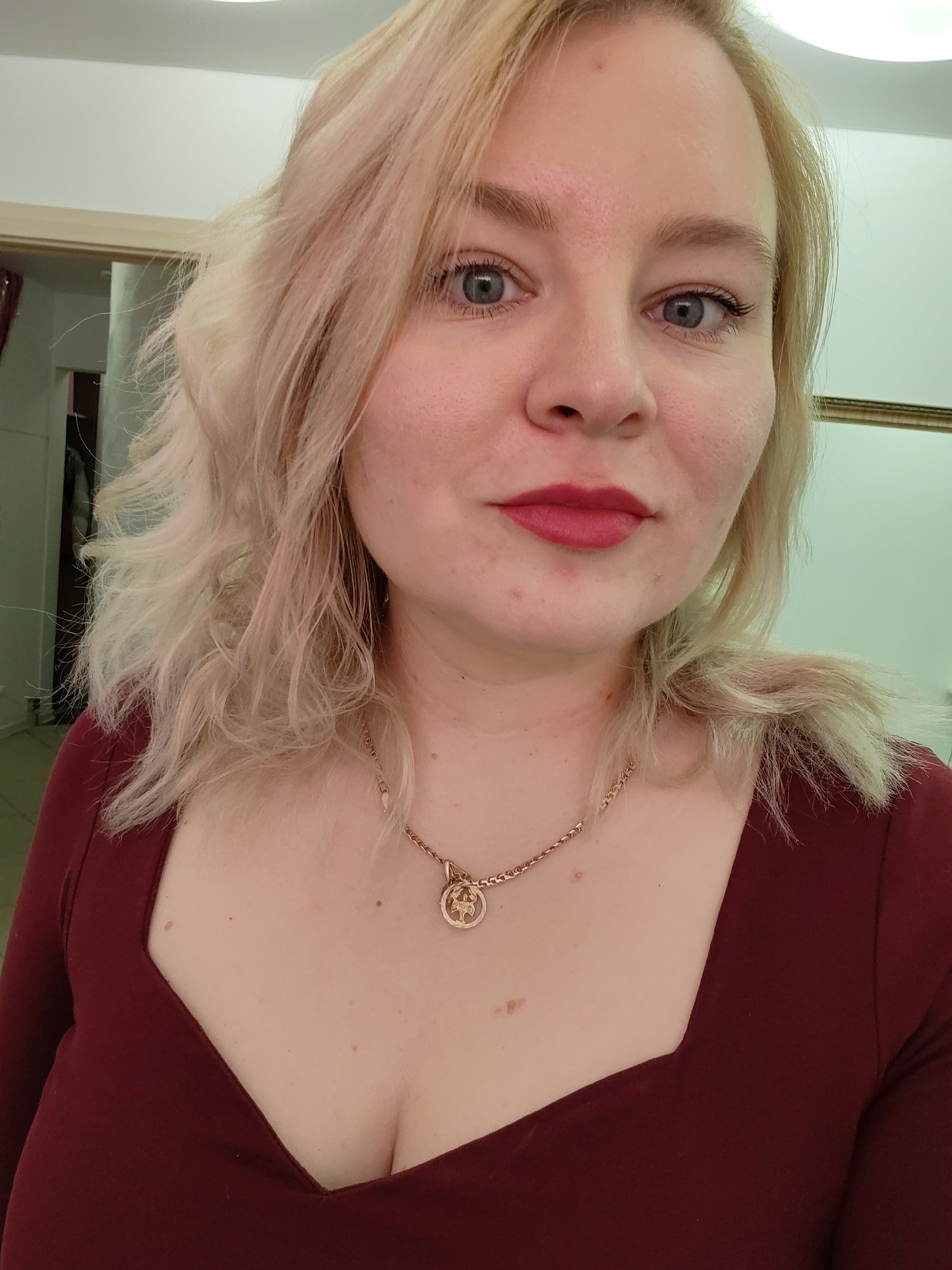 A portrait photograph features an adult woman with fair skin, looking directly at the camera. She has shoulder-length, wavy, light blonde hair and light blue eyes. Her eyebrows are light, and she wears bright red lipstick on her full lips. Her complexion shows some natural skin variations, including moles and minor blemishes on her face and neck, and faint stubble is visible around her mouth.
She is dressed in a dark red, long-sleeved top with a broad, square neckline. A gold-colored chain necklace featuring a circular pendant, which appears to depict a stylized tree or abstract design, is worn around her neck.
The background indicates an indoor setting with light-colored walls. On the left, part of a doorframe or archway is visible, while on the right, a sliver of a reflective surface or framed object can be seen. The scene is lit by overhead artificial lighting.
The woman maintains a neutral and calm expression, with a direct gaze, suggesting she is posing for the photograph. There is no discernible text in the image. This photo was taken in Izhevsk, Russia.