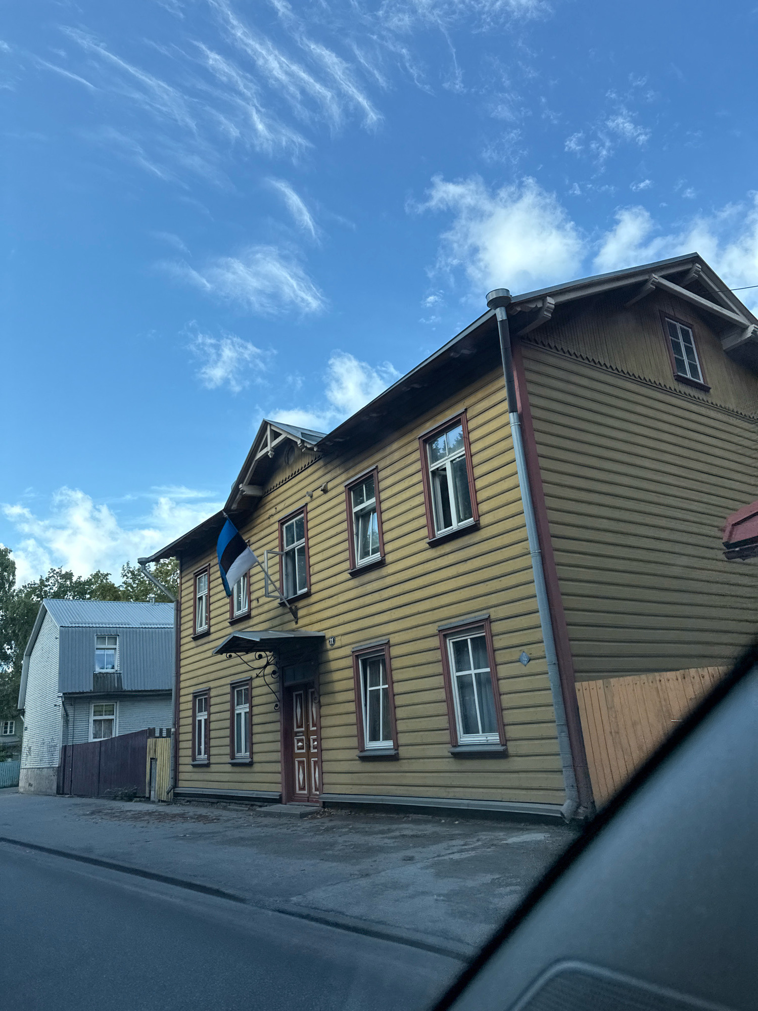 The image captures a street scene in Tallinn, Estonia, with a prominent wooden house painted yellow and adorned with the Estonian flag. The house features multiple windows with dark red trim and a dark red door. To the left of the yellow house, a modern-looking building with a silver corrugated metal facade is visible, separated by a dark brown fence. The street in the foreground is paved and appears to be asphalt. The sky above is blue with wispy white clouds, indicating daytime. The photograph is taken from the perspective of a vehicle, as suggested by the blurred car interior in the lower right corner of the frame. The overall impression is a snapshot of a residential area in Tallinn on a clear day.