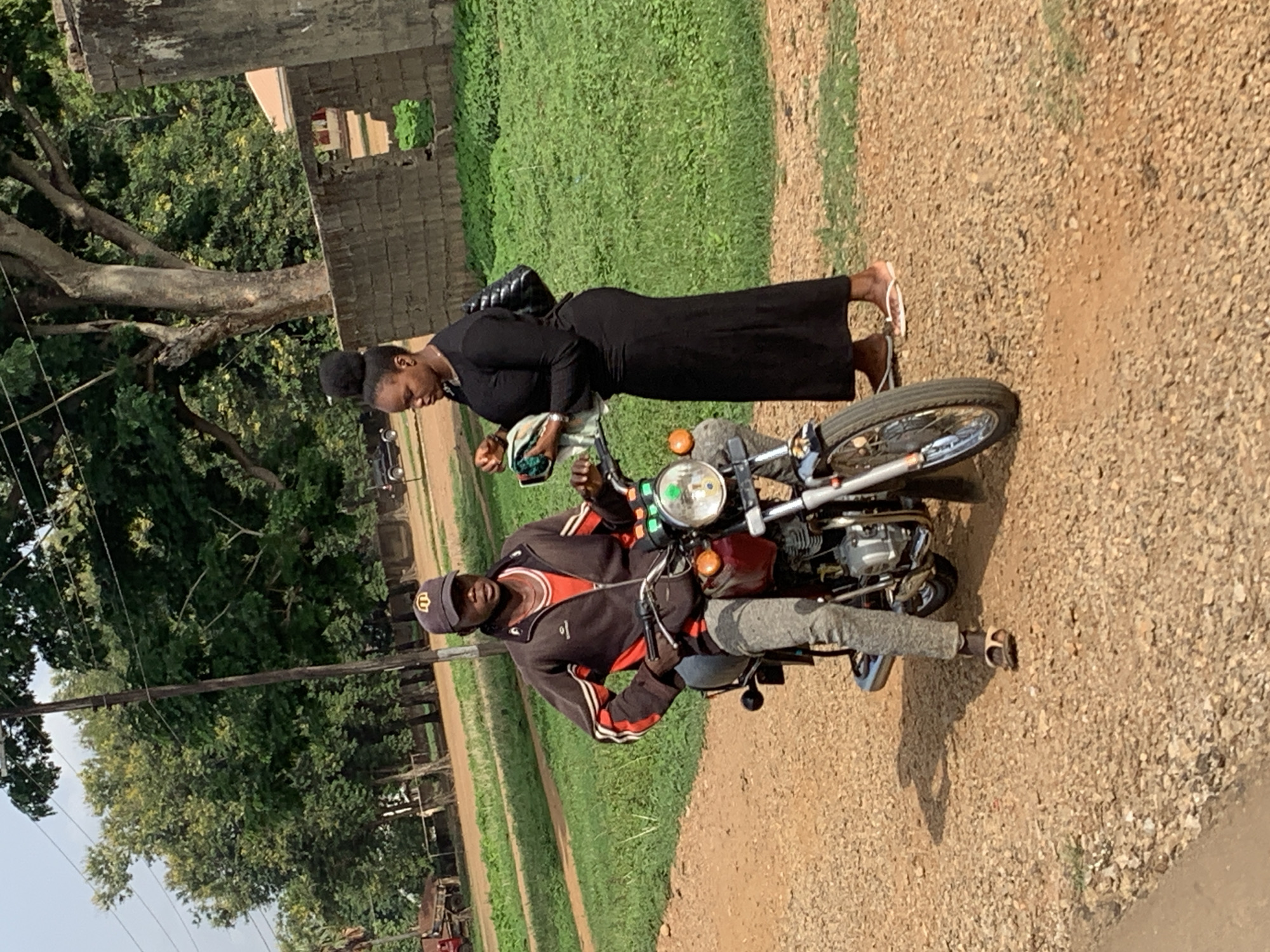 The image captures a scene in Agban, Nigeria, featuring a man seated on a motorcycle and a woman standing beside it. The man is wearing a dark-colored jacket with red and white stripes, grey pants, a cap, and sandals. The woman is dressed in a long black dress and sandals. She has a bag over her shoulder. The motorcycle is red with chrome details. They are positioned on a gravel road next to a grassy area. The background includes trees, a building, and utility poles.