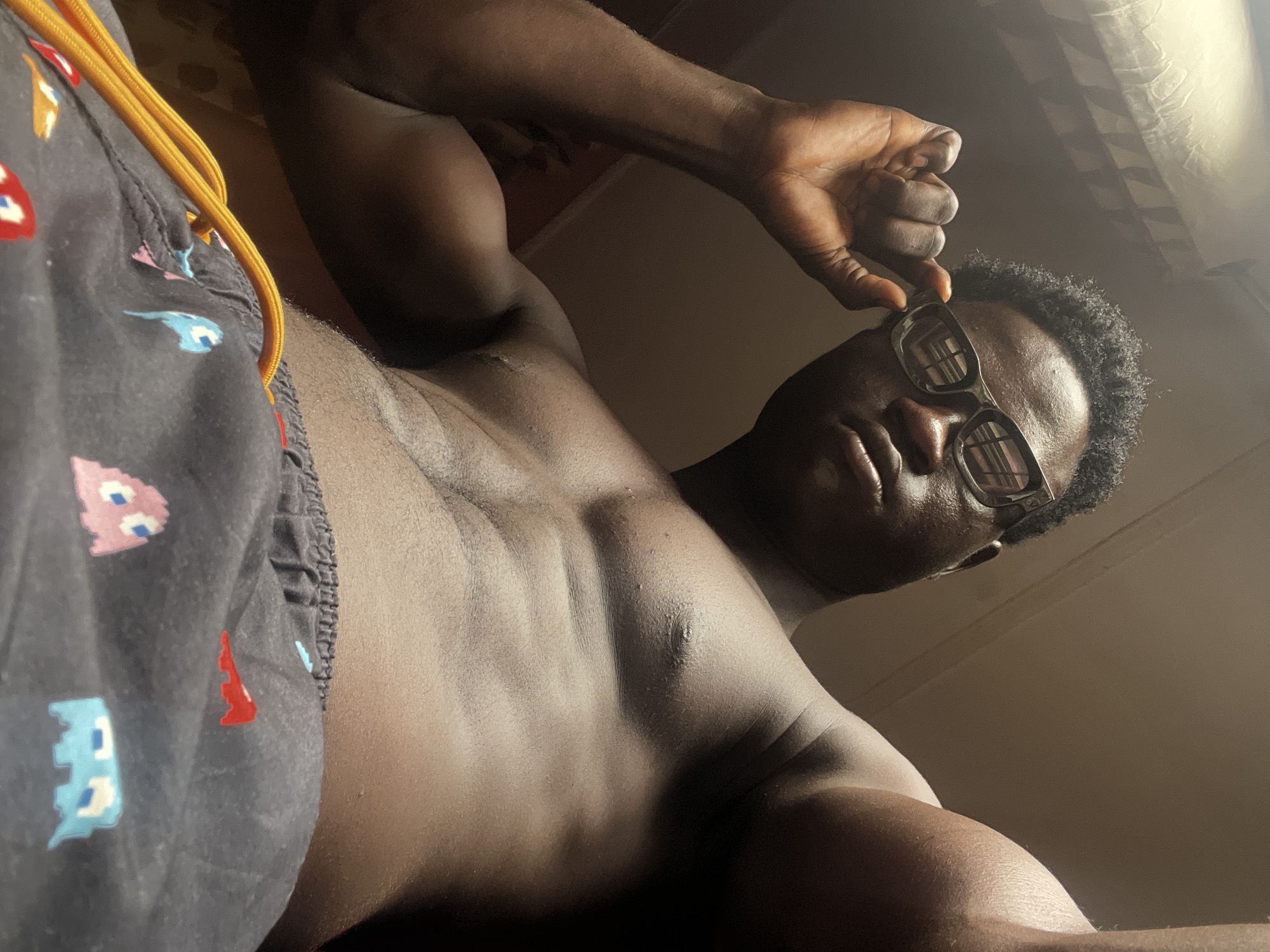 A low-angle shot shows a shirtless dark-skinned man in eyeglasses adjusting his glasses with his right hand. He is wearing shorts with Pac-Man characters printed on them. The background is blurry, but appears to be an indoor space, perhaps a bedroom. Location cannot be determined to be Mosafejo, Nigeria.