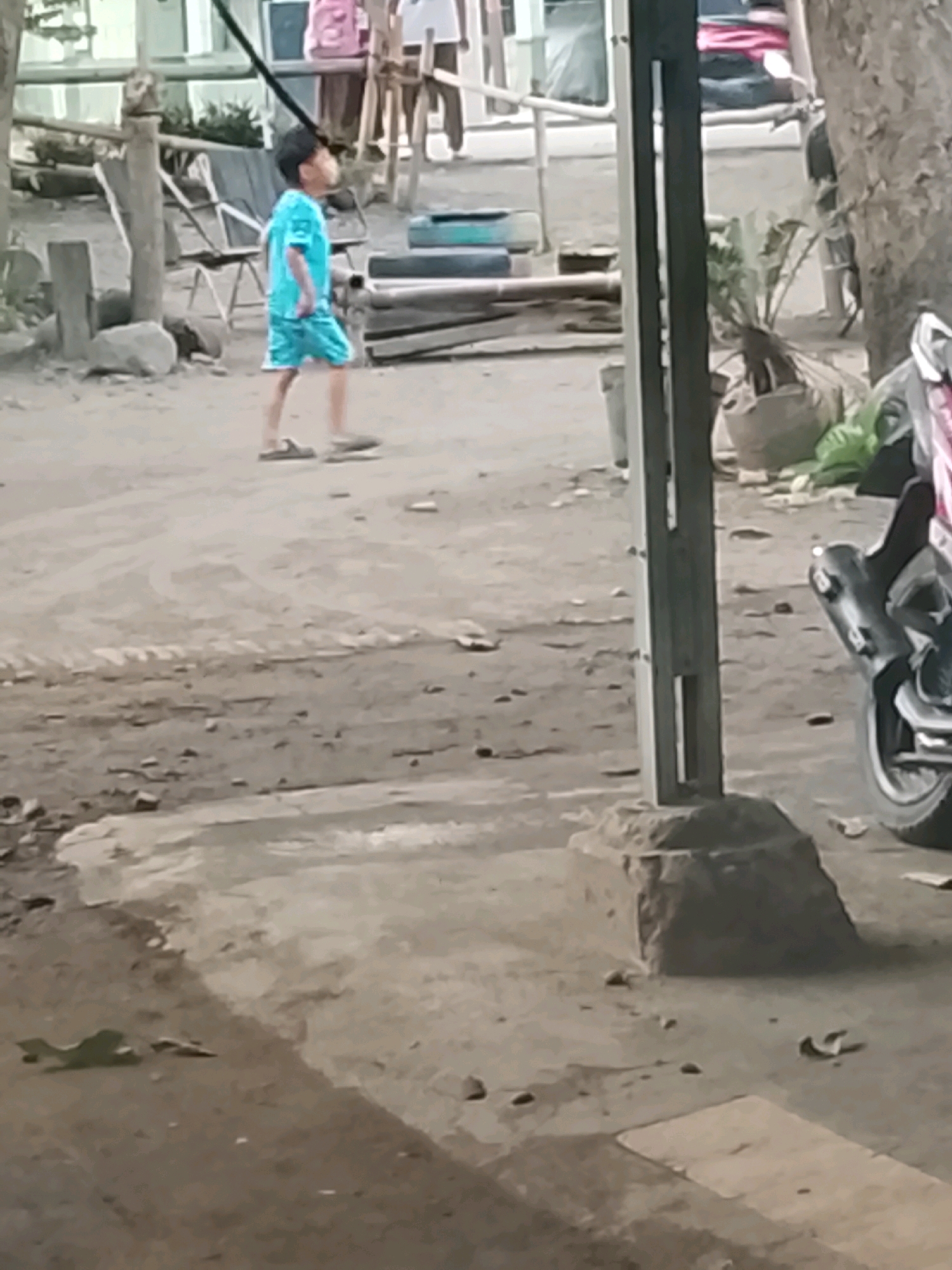 A young boy wearing a turquoise t-shirt and shorts walks on a dirt path. He is facing to the left of the frame, with his head turned to the right. In the background, there are some wooden structures, chairs, and a motorcycle parked on the right side. The ground is uneven and covered in dirt and debris. The overall impression is of an outdoor, possibly rural, setting in Ujungrusi, Indonesia, where everyday activities are taking place.