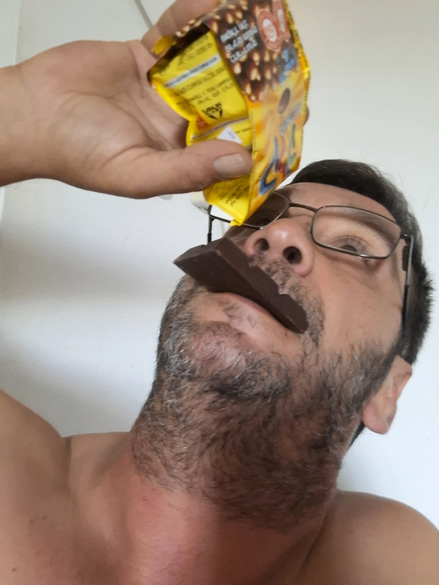 A close-up, low-angle shot shows a man's face as he holds a piece of chocolate in his mouth. He is wearing glasses and has a beard. The man's hand, holding a crumpled yellow wrapper, is visible in the upper left corner of the image. The background is a plain white wall. The image appears to have been taken indoors in Yerocham, Israel.
