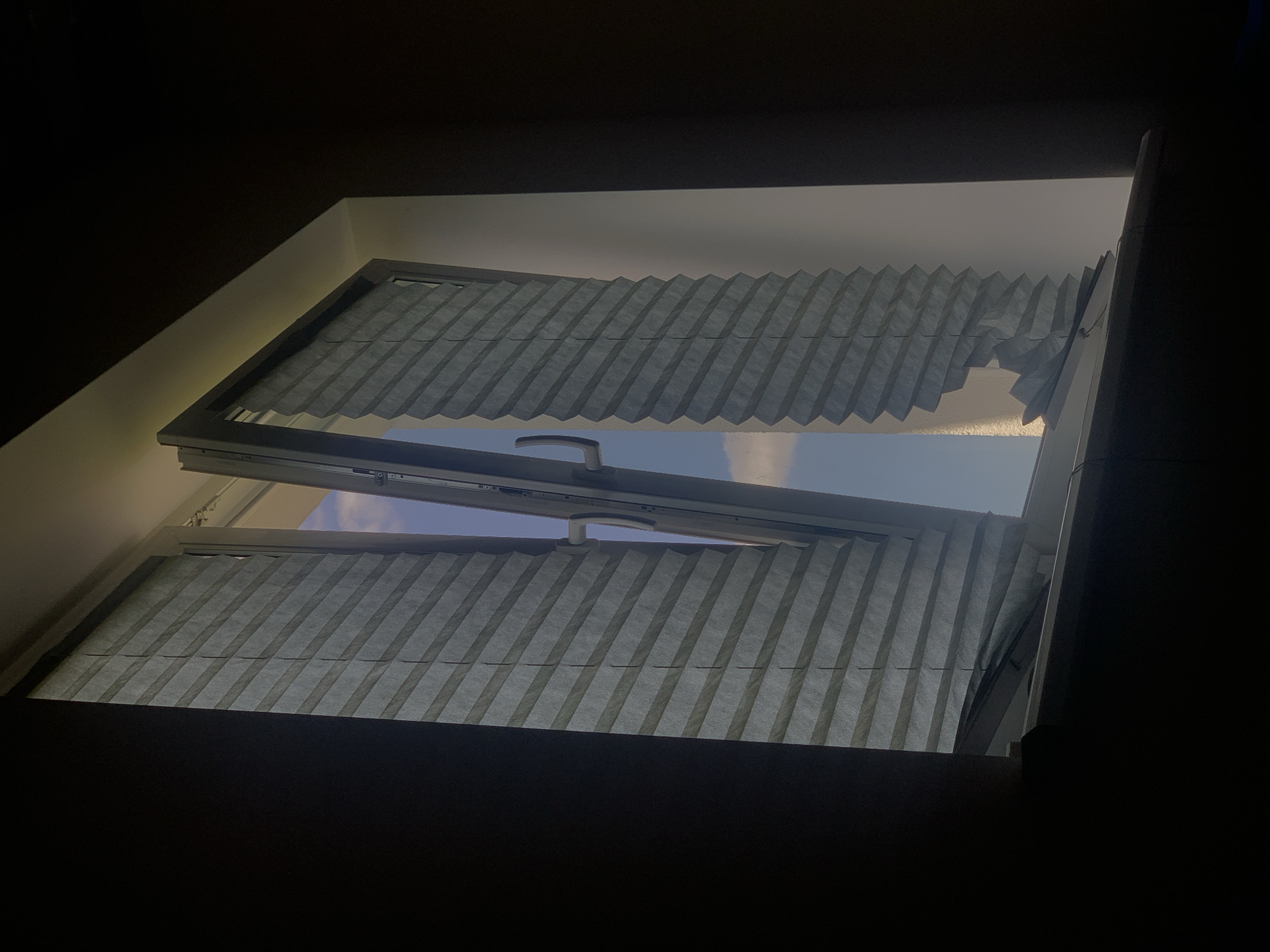 An interior view of an opened window with pleated blinds, showing a glimpse of the sky. The left blind is mostly intact, while the right blind is torn at the bottom. The window frame is light gray, and a silver handle is visible on the right side of the frame. The surrounding walls are dark, suggesting it might be nighttime or the room is dimly lit. The scene is set on a windowsill in a residential building located in Minsk, Belarus. No people or animals are present.