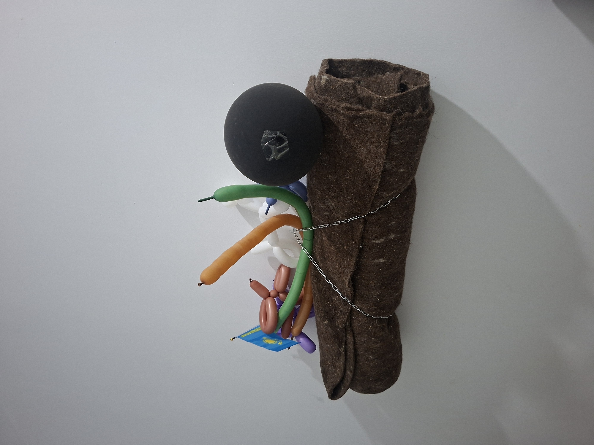 An indoor photograph shows a peculiar art installation against a plain white wall. The installation consists of several disparate elements: a large black sphere with a square of clear tape affixed to its surface, a collection of brightly colored balloon animals in various shapes and sizes, a small blue flag with a design visible, and a rolled-up brown blanket secured with a silver chain. The flag appears to be under the balloon animals. The composition is unusual and abstract. No people or specific environmental elements are identifiable beyond the neutral wall. There is no indication of Kokshetau, Kazakhstan, in the visual data provided.