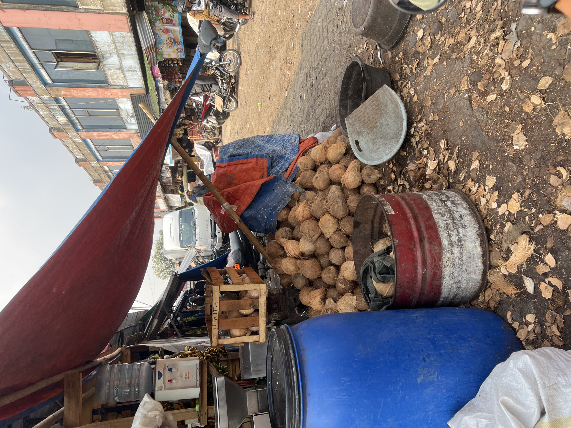 The image depicts an outdoor market scene in Citeureup, Indonesia, characterized by a bustling atmosphere and a display of fresh produce. A prominent red and blue tarp stretches over the scene, providing shade. In the foreground, a large blue barrel sits beside a red and white striped metal barrel filled with coconuts and green fabric. A substantial pile of coconuts is also visible on the ground, with scattered coconut husks. A wooden crate labeled "HRITA" contains more coconuts. Behind these, a water dispenser with two spigots is situated near a cluster of bananas. The background reveals a row of two-story buildings with numerous windows, some open. Several motorcycles and a white Isuzu truck are parked in the area, suggesting ongoing commerce. A few individuals are present, including a person on a motorcycle in the lower right corner. The ground is dirt and scattered debris, typical of an outdoor market environment.