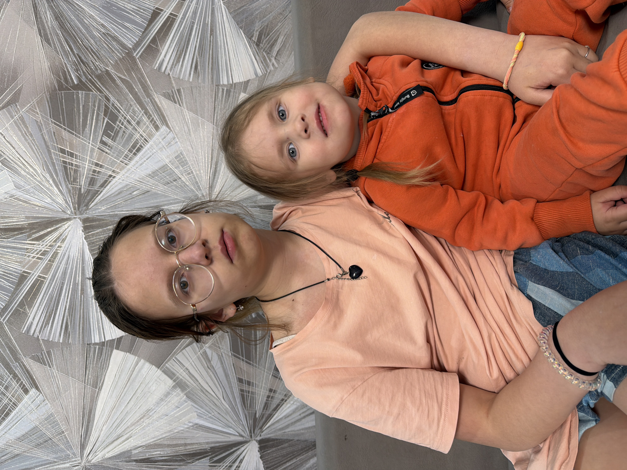 The image captures two individuals: a young girl and an adolescent girl. The young girl is dressed in an orange hooded sweatshirt. Her hair is light blonde and styled into pigtails. The adolescent girl is wearing a peach-colored t-shirt and has round glasses. She has dark hair pulled back from her face. A necklace with a dark stone is visible around her neck. The girls appear to be reclining or lying down on a gray couch. The background features a wall with a repeating geometric pattern in shades of gray and white. The location is Chaikovsky, Russia.