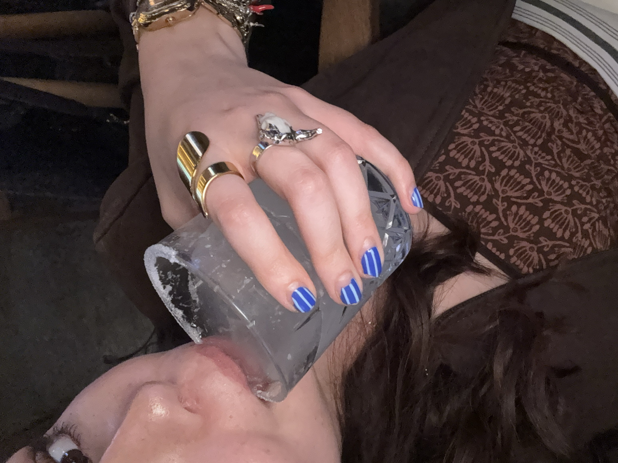A close-up shot from an unusual angle shows a person drinking from a clear, ice-filled glass. The person has dark, wavy hair and is wearing a brown top over a floral-patterned fabric, possibly a dress or bedding. The nails on the hand holding the glass are painted blue with white stripes. The hand also sports several rings, including a large gold ring, a sculpted silver ring with white detail, and a wide, abstract gold ring. A gold wristwatch is visible on the wrist. The background is dark and indistinct. The location is unknown, and there is no indication that it is Mexico City, Mexico.