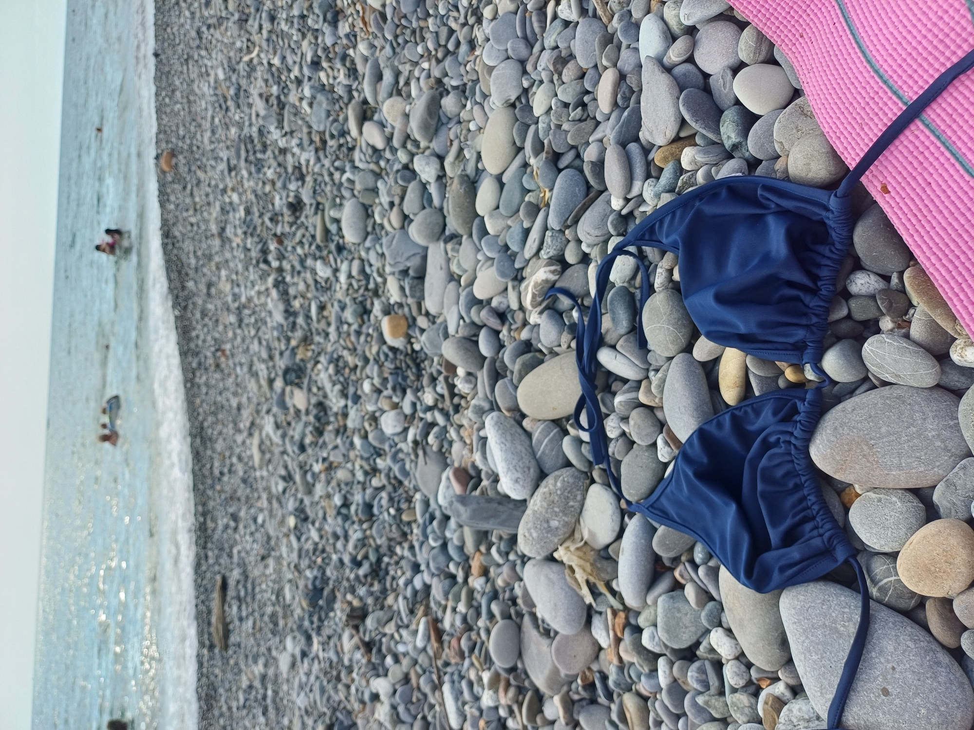 A bikini top and a pink straw hat rest on a pebble beach in Sochi, Russia. The bikini top is navy blue and made of a shiny fabric, with thin straps and gathered fabric on the cups. The hat is pink and woven, with a darker pink trim. The beach is covered in smooth, gray, and white pebbles of various sizes. To the left of the beach, the clear turquoise water of the Black Sea is visible, with small waves gently lapping at the shore. Two people are partially visible in the water, appearing to be swimming or wading. The sun is shining, casting a gentle sparkle on the water. The scene suggests a relaxing day at the beach, possibly during the daytime or late afternoon given the soft light. The location is identifiable as Sochi, Russia, known for its pebble beaches and the Black Sea.