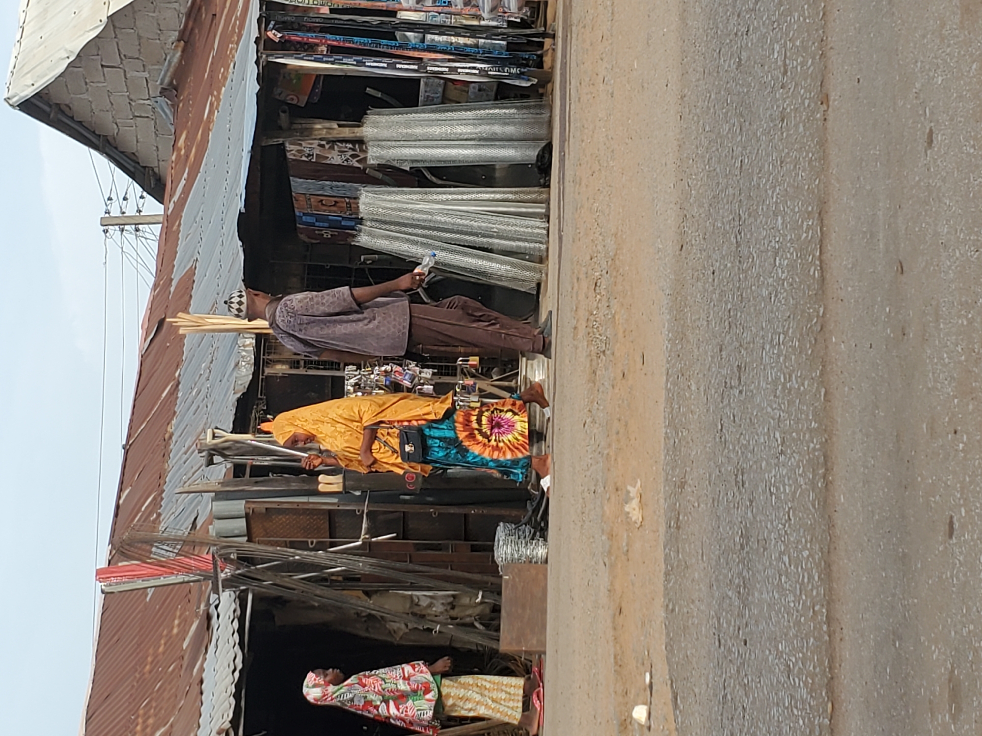 This upright image depicts a vibrant street scene, likely in a market or commercial area, in Mararaba, Nigeria, under clear, sunny daytime conditions.
In the foreground, a dusty, unpaved road or pathway extends across the right side of the frame. On the left, a series of makeshift stalls or small shops with corrugated iron roofs line the street, showcasing various goods.
Several people are visible in the scene. A man, wearing a light-colored long-sleeved tunic, dark trousers, and a white patterned cap, stands holding a plastic bottle. He is positioned near stacks of what appear to be building materials, possibly aluminum profiles or pipes, wrapped in protective plastic. Further back, another person is seen wearing a bright yellow head covering and top, complemented by a colorful tie-dye patterned skirt featuring shades of blue, orange, and pink. This individual also has a black bag slung over their shoulder. Closer to the foreground and lower in the frame, a third person wears a white head covering with a colorful floral design and a matching top, over a striped skirt.
The stalls are filled with merchandise. Shelves display various items that could be hardware, household goods, or electronics. Prominent stacks of metal rods or poles are visible, suggesting the sale of construction materials. Above the stalls, numerous power lines and cables are strung between wooden poles.
The overall atmosphere suggests an active commercial environment. A partially visible text "OMO HOHO" can be deciphered on a package displayed in one of the upper stalls.