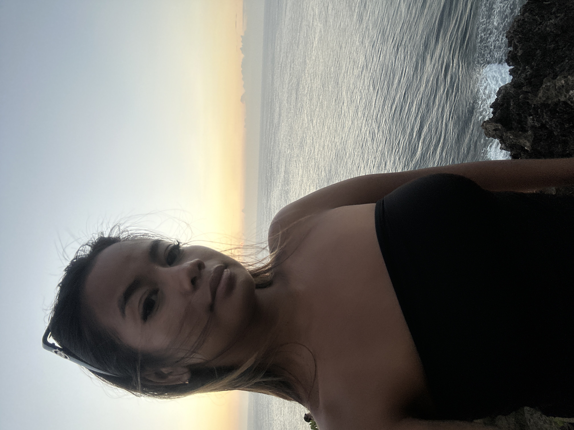 This media file is a portrait of a young woman taken in Kuta Selatan, Indonesia. The woman is positioned in the foreground, with a dusky sky and the ocean as her backdrop. She is wearing a black strapless top and sunglasses perched on her head. The sun appears to be setting, casting a warm, golden glow on the horizon, which gradually fades into a lighter sky. The ocean is calm with gentle waves, and a rocky outcrop is visible to the right, suggesting a coastal environment. The woman's hair is slightly windswept, adding to the natural and serene feel of the image. Her expression is relaxed, and the overall mood is peaceful and contemplative.