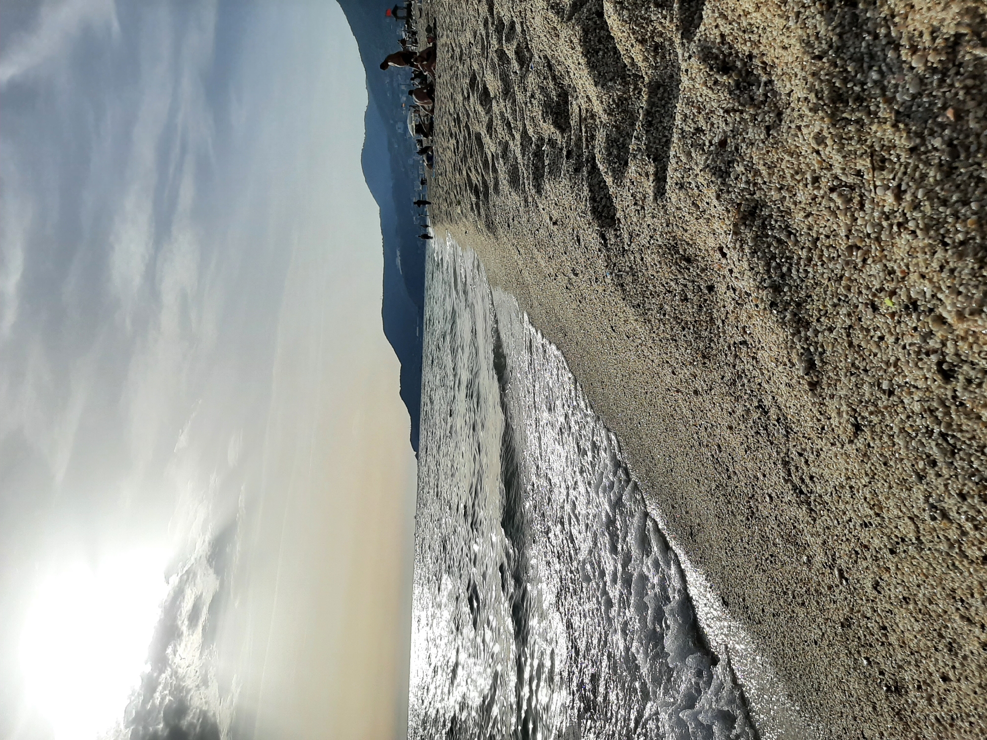 The image is a beach scene in Alanya, Turkey. The foreground consists of wet pebbles merging into calm, shallow seawater. Sunlight reflects off the water's surface. The background shows a mountain range and the hazy sky. In the distance, on the beach, there are sparse figures of people under umbrellas.