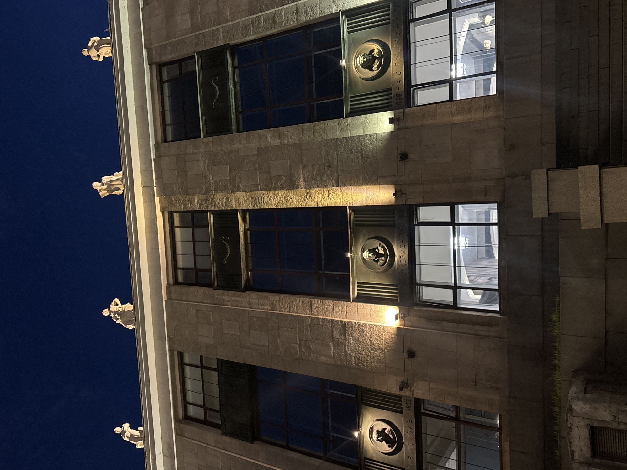 This is a nighttime, low-angle exterior shot of the exterior of a stone building in Moscow, Russia. The building has large, dark windows and is adorned with bas-relief portraits of historical figures, specifically Nikolai Gogol and Alexander Pushkin, along with dates of their lives. Statues of figures in classical attire are positioned on the rooftop. The building's facade is illuminated by artificial lights.