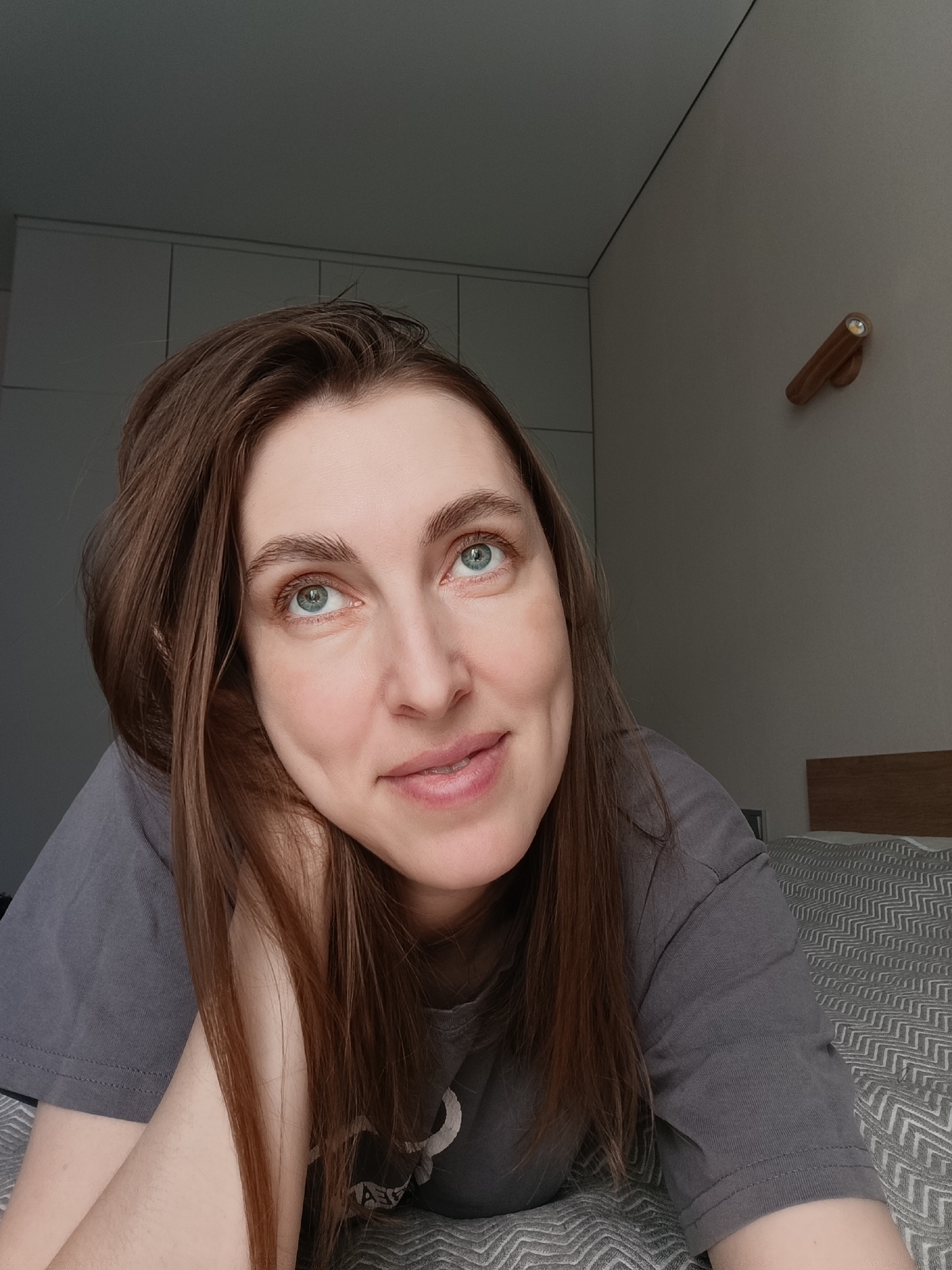 A woman with long brown hair and light blue eyes is lying on a bed with a grey and white patterned blanket. She is wearing a grey t-shirt and resting her head on her hand. Behind her is a light grey wall with a wooden fixture attached. To the left is a built-in closet with white doors. The photograph appears to have been taken indoors, possibly in a bedroom, in the city of Chaikovsky, Russia.