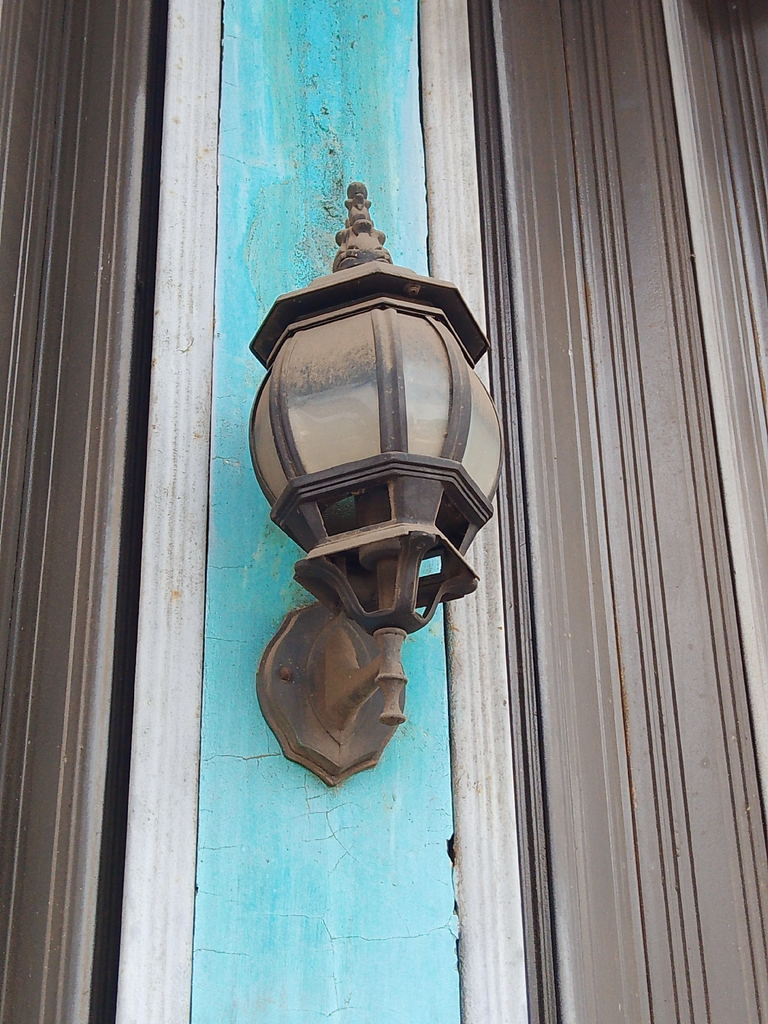 A close-up, low-angle outdoor shot captures a vintage wall-mounted lantern affixed to a cracked, turquoise-painted surface. The lantern, made of dark metal with glass panes, appears weathered and dusty, suggesting age and exposure to the elements. It is mounted on a decorative metal bracket, which in turn is attached to the wall with a visible screw. The background consists of vertical corrugated metal panels, one silver-gray and the other dark gray, creating a textured and industrial feel. The context of Potroyudan, Indonesia, suggests a potentially older building or historical area.