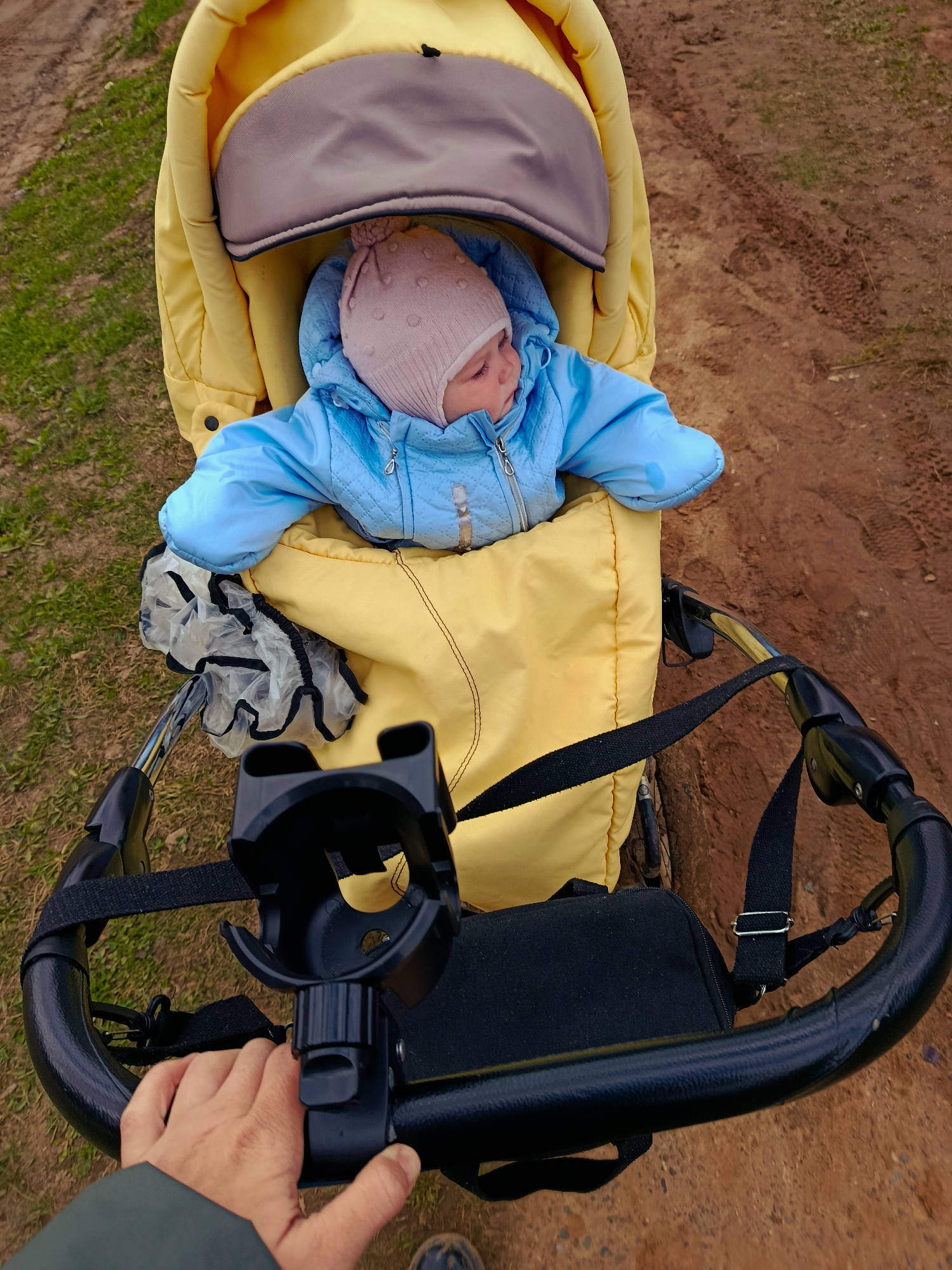 A high-angle, full shot depicts a baby in a yellow stroller being pushed by a person on a dirt path.
The baby wears a pink hat and a blue jacket, nestled within the yellow stroller's padding, its head tilted slightly to the right. The stroller features a gray hood and a black handle held by a hand wearing a green sleeve. A black bag and a cup holder are attached to the stroller.
The surroundings consist of a dirt path with patches of green grass along the edges.
The location may be Фоки, Russia.
