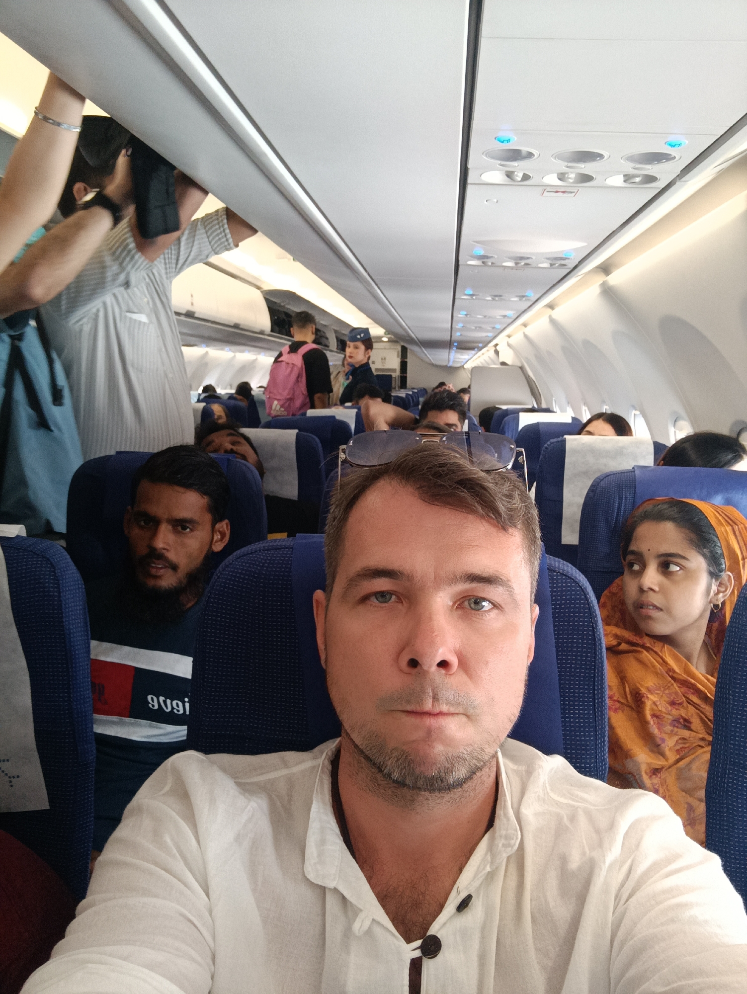 The image is a selfie taken inside an airplane cabin, capturing passengers during what appears to be boarding or deplaning. The main subject, a man with light skin, short light brown hair, and stubble, wears a white collared shirt and looks directly into the camera with a neutral expression. Sunglasses are perched on his head.
Behind him, to his left, a man with dark skin and a beard wears a dark t-shirt with a white and red stripe, showing the reversed text "9U9i" (likely a reflected or mirrored brand logo, possibly IndiGo). He looks towards the left. To the main subject's right, a woman with dark skin, wearing an orange patterned headscarf and traditional attire, looks to her right with a slightly pensive expression.
The cabin features rows of blue fabric seats with white headrest covers. Overhead bins are visible, with some open and luggage being stored or retrieved. In the aisle, other passengers are present, including one person reaching into an overhead bin, another with a pink backpack, and a flight attendant in a dark uniform with a blue scarf. The ceiling shows lights and air vents. The artificial interior lighting suggests the event takes place during the day. The visible text and the appearance of the passengers are consistent with the provided location context of Anneshvara, India, suggesting an Indian airline or travel within the region.