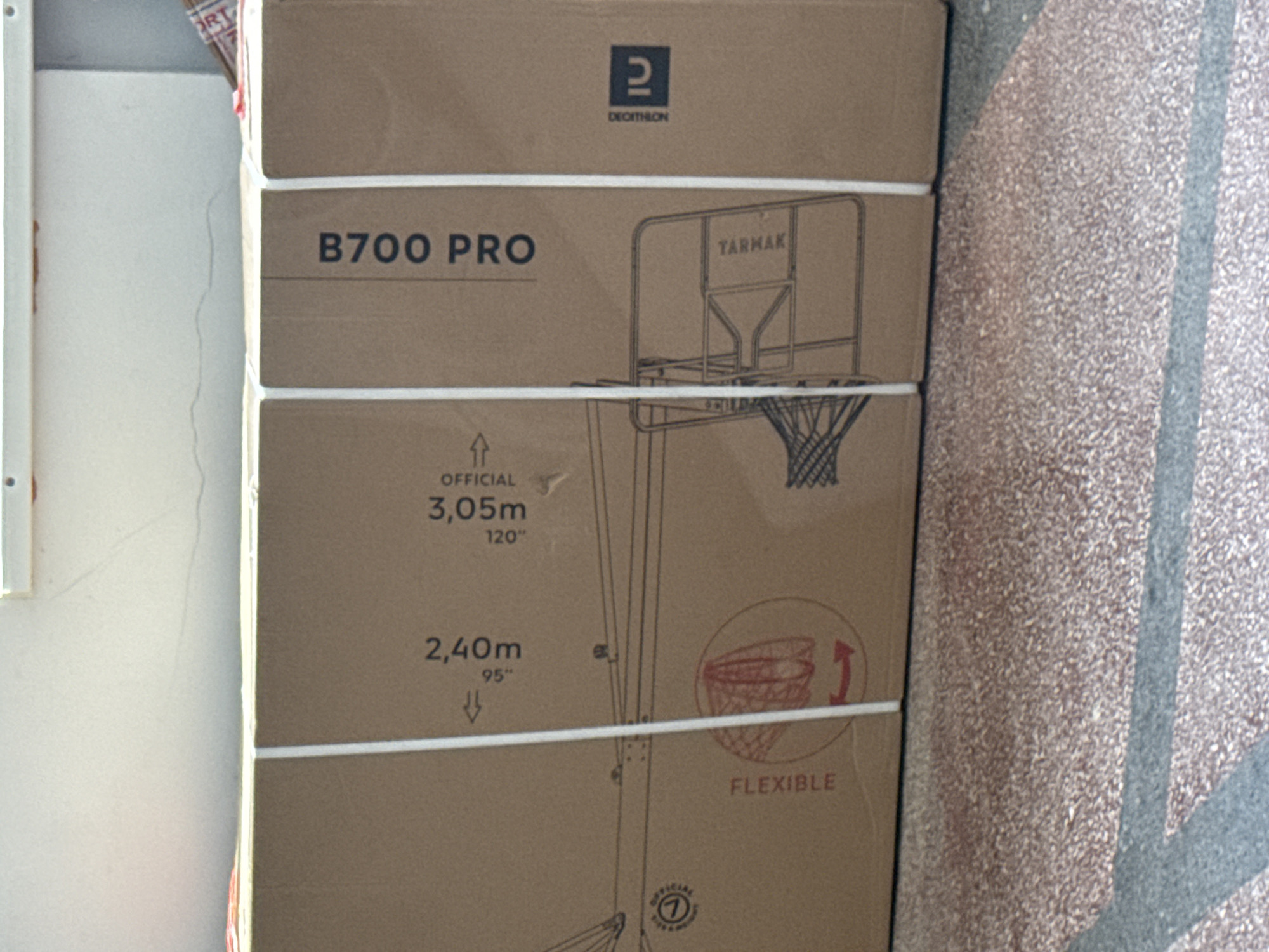 The image captures a large cardboard box, likely containing a basketball hoop, standing upright against a plain white wall. To the right, a textured surface, possibly a floor or wall with a speckled pattern in brown and grey tones, is visible. The box is secured by two horizontal white plastic straps.
The front of the cardboard box features several black printed graphics and text. At the top, the "2 DECATHLON" logo is clearly visible, although "DECATHLON" is slightly obscured and was OCR'd as "DECITHION". Below this, the product model "B700 PRO" is printed. An illustration of a basketball hoop with a backboard displays the brand name "TARMAK" on the backboard.
Further details printed on the box include height specifications: "OFFICIAL 3,05m 120"" and a lower setting of "2,40m 95"", each accompanied by an arrow indicating adjustability. An illustration depicts a basketball net with a circular arrow, labeled "FLEXIBLE", suggesting a flexible rim. The word "OFFICIAL" appears again near a circled number "7" logo. A partial word "ORT" from another object or packaging is visible in the top left corner.
The scene appears to be an indoor or semi-indoor environment, possibly a storage area or hallway, with general ambient lighting. There are no people or discernible activities. The image provides a detailed view of the product packaging. This media file was captured in Varna, Bulgaria.