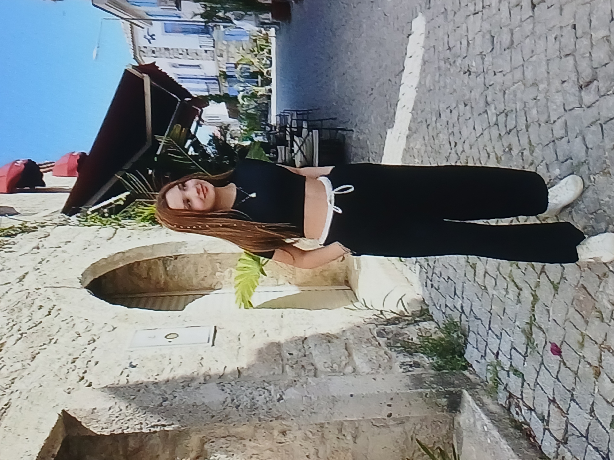 A young woman with long, light brown hair, styled with braids and decorative beads, stands on a cobblestone street. She is wearing a black crop top, black wide-leg pants with a white drawstring waistband, and white sneakers. Her right hand is in her pocket, and her left arm is bent at the elbow. She is looking to her right with a slight smile. Behind her, to the left, is a stone building with an arched opening. To the right, the street continues, lined with buildings, awnings, and outdoor seating for a cafe. The scene appears to be in an urban setting, possibly a historic district, with an overall bright and sunny atmosphere. The image does not contain any elements that suggest it is located in Dnipro, Ukraine.