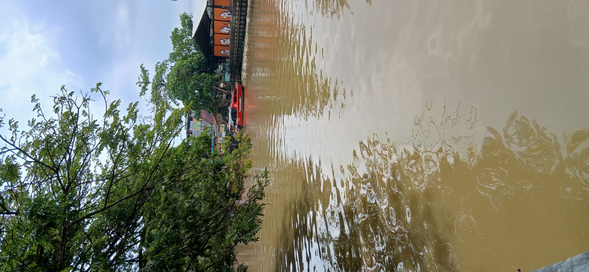 A still, wide shot captures a muddy brown river flowing through the city of Samarinda, Indonesia. Lush green trees overhang the left side of the frame, their branches casting dappled shadows on the water's surface. On the right bank, a bright red boat with white lettering is docked. Behind it, a low-slung building with an orange, graffiti-covered wall and a corrugated metal roof stands. The sky above is overcast with gray clouds. The scene is quiet and still, with no visible people.