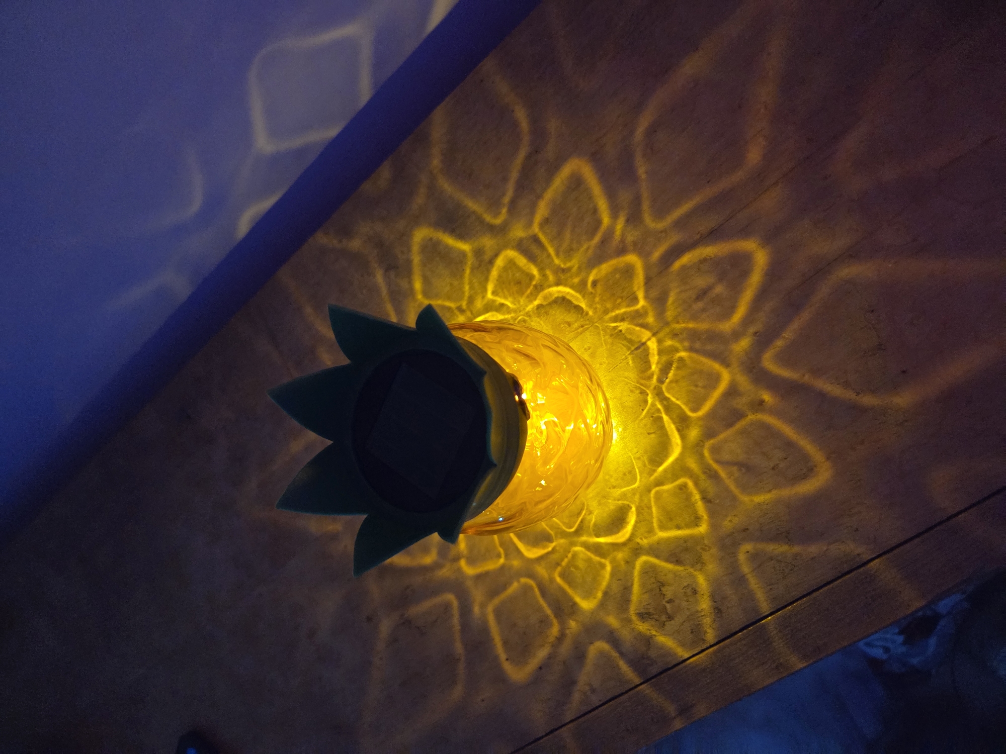 A top-down view shows a pineapple-shaped outdoor solar light sitting on a wooden surface. The light emits a bright yellow glow, projecting a patterned light design onto the surface. The light's top is shaped like the pineapple's green crown. The background is partially visible, with a blue-tinged wall or surface on the left side of the image. There is no discernible location information related to Cherkovitsa, Bulgaria.