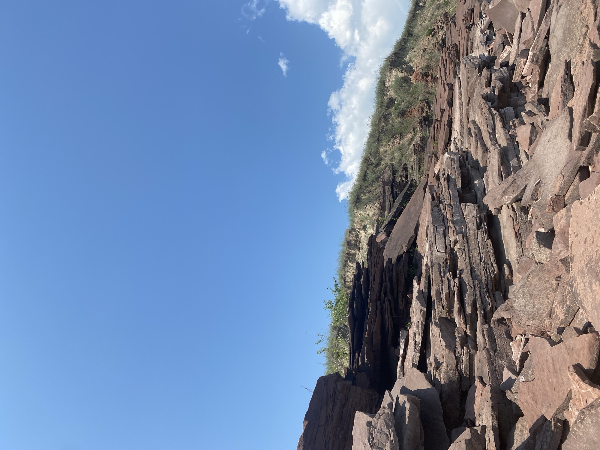 The image shows a rocky, uneven terrain with large, flat, reddish-brown stones stacked and scattered across the foreground and midground. Behind the rocks, a steep, grassy embankment rises, topped by a clear blue sky with a few wispy white clouds. The environment appears natural and undeveloped, suggesting an outdoor, possibly remote location. The scene is devoid of any human presence or activity. The location is identified as the city of Ust'-Erba, Russia, implying a natural landscape within or near this area.