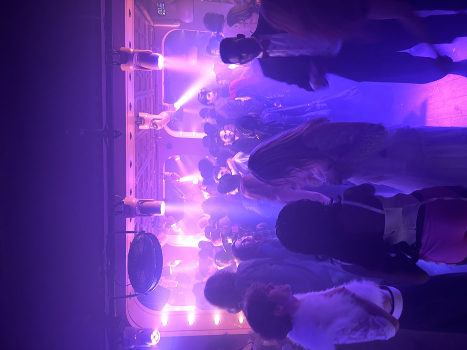 A crowded nightclub scene is illuminated by blue and purple lighting. Robotic arms with spotlights shine over the crowd. People in varying outfits fill the space. A fan is visible on the side, adding to the energetic atmosphere. The image is taken from a slightly elevated perspective, capturing the density of the crowd and the dynamic lighting effects.