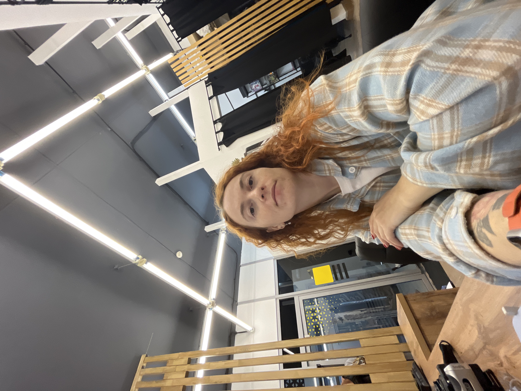 A woman with red hair is in a building with fluorescent lighting, wood slats, and a window. The building is located in Chaikovsky, Russia. The woman is wearing a plaid shirt. There is camera equipment on a wooden surface in front of her. The background includes a black curtain and a gray ceiling.