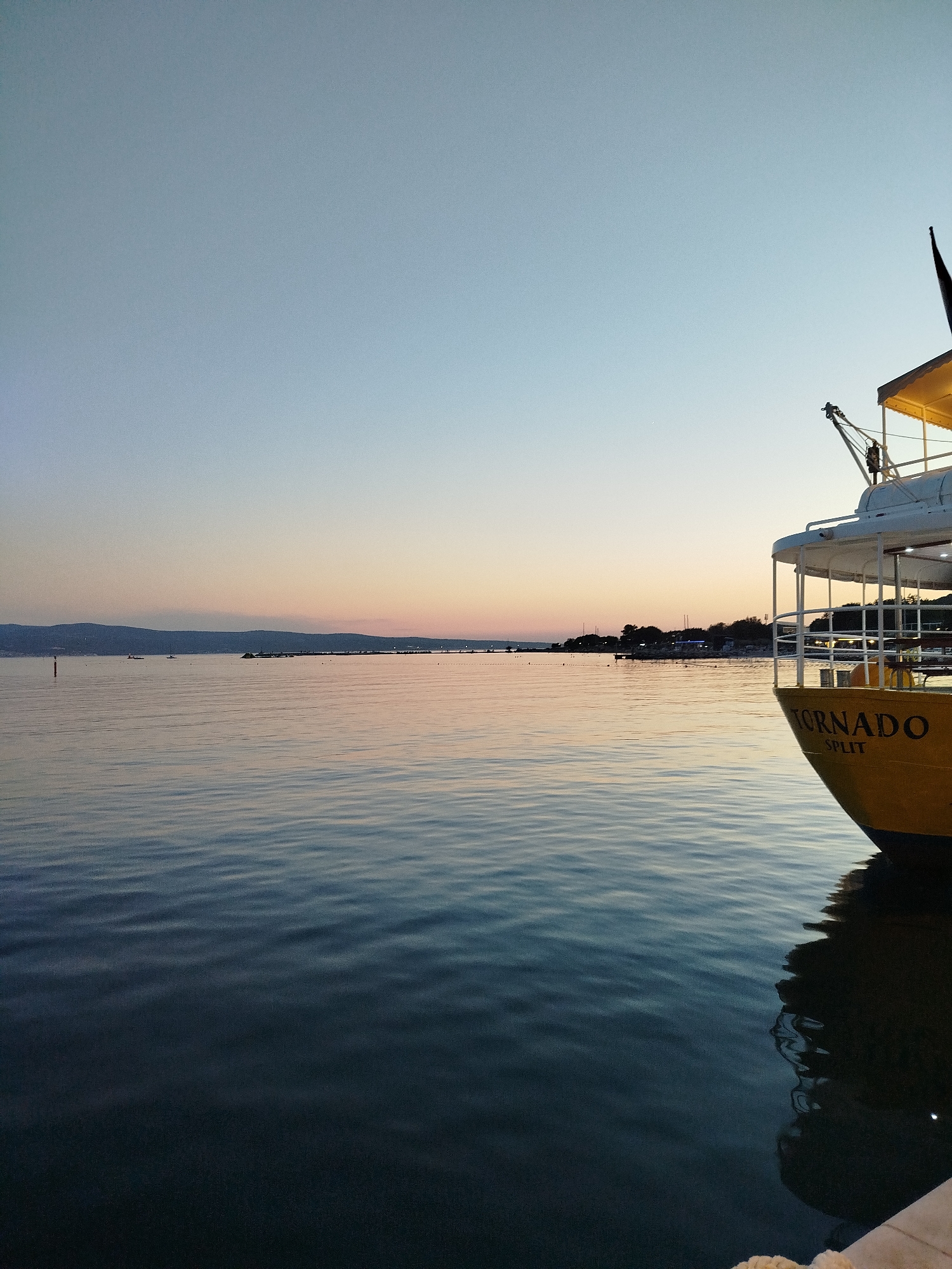 An outdoor, eye-level, medium shot captures a serene coastal scene at dusk. The sky transitions from a pale blue at the top to a soft gradient of pink and orange near the horizon. The calm water, reflecting the sky's colors, fills the foreground and mid-ground, with subtle ripples creating a textured surface. On the right, the stern of a yellow boat, identified by the words "TORNADO SPLIT" in black lettering, is prominently featured, its white upper deck structure partially visible. A crane extends from the boat's deck. In the distance, a dark, low-lying landmass with a few scattered lights suggests the presence of a coastline or a town. The overall ambiance is peaceful and tranquil, indicative of the end of the day.