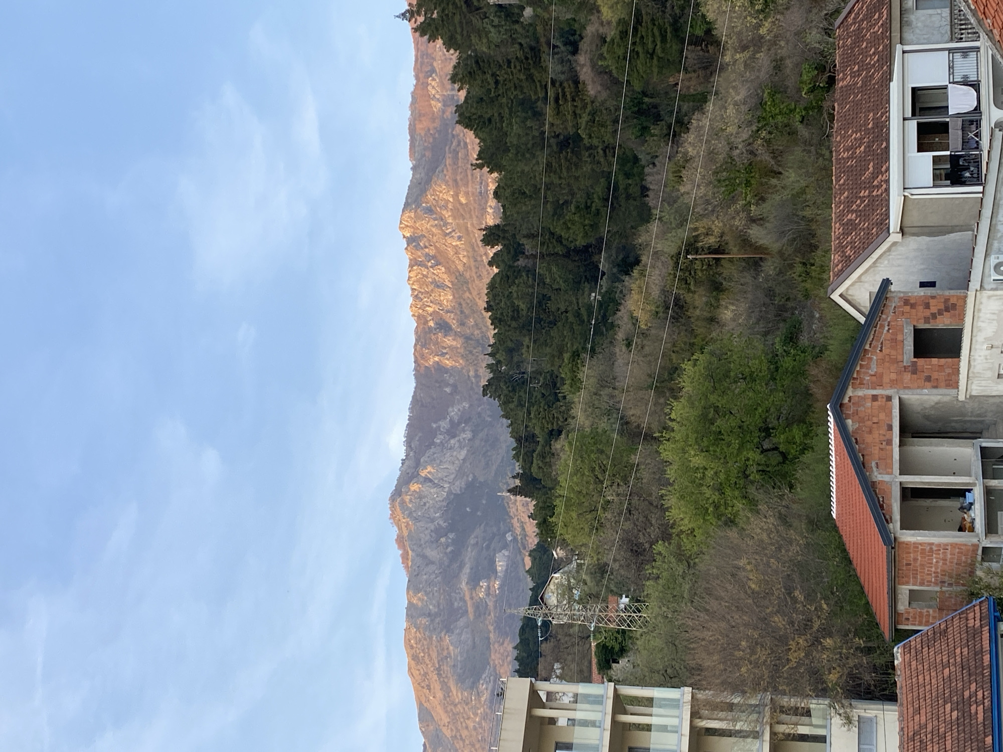 The image depicts a view in Budva, Montenegro, showcasing a mountain range bathed in the warm light of either sunrise or sunset. The sky is a pale blue with subtle clouds. The mountain slopes are partially covered in dense forest, with some areas revealing rocky surfaces. Power lines stretch across the foreground and midground, adding a human-made element to the natural landscape. Buildings, including residential structures in various stages of construction or completion, are visible in the lower right corner. A multi-story building with glass balconies is situated towards the bottom of the image. The scene appears to be taken from a high vantage point, looking out towards the mountain and the surrounding area.