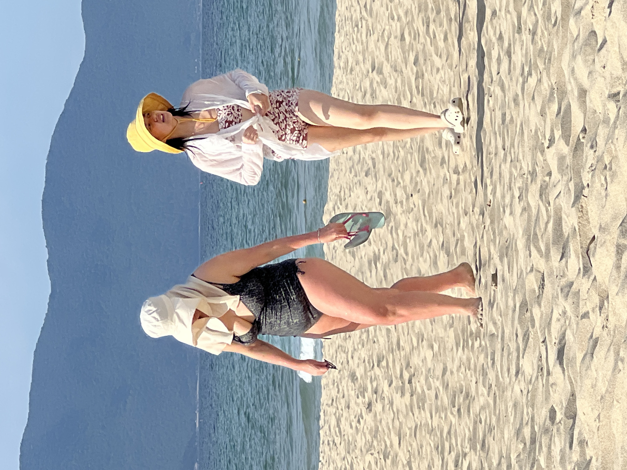 Two people are on a sandy beach in Đà Nẵng, Vietnam. The image is oriented vertically.
In the upper portion of the image, a person wearing a yellow sun hat, a white sheer cover-up, and a patterned bikini top is standing. They are holding the edges of the cover-up. Their mouth is open as if speaking or laughing. White shoes are visible on their feet.
In the lower portion of the image, another person is bent over, wearing a dark one-piece swimsuit and a light-colored sun hat. They are holding a pair of flip-flops in one hand and a phone in the other. Their legs are extended behind them.
The background shows the calm, blue ocean meeting the shore, with a large, dark mountain range in the distance under a clear blue sky. The sand is light-colored and has ripples and shadows, indicating it's likely daytime with bright sunlight. The overall atmosphere appears relaxed and summery.