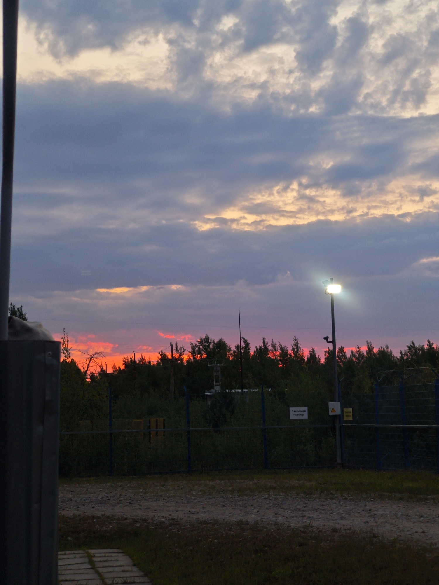 This detailed image captures a twilight scene, likely a sunset, over an industrial or utility area. The sky dominates the upper portion, displaying a palette of dark blue-grey clouds interspersed with bands of soft orange and beige, indicative of fading daylight. Below this, a striking, intense red-orange glow from the setting or rising sun is visible just above the tree line, creating a dramatic contrast with the darker clouds.
In the midground, a dense line of dark green trees forms a silhouette against the vibrant red sky. A tall, dark grey utility pole stands centrally on the right, equipped with a brightly illuminated floodlight that emits a strong white glare. Below the floodlight, a smaller, dark, unidentifiable object, possibly a camera or sensor, is attached to the pole.
Stretching horizontally across the lower midground is a dark blue chain-link fence, topped with what appears to be barbed wire or a similar deterrent. Attached to the fence or its posts are several signs: a white rectangular sign on the left with black Cyrillic text reading "Запретная граница" (Forbidden boundary/border), a yellow triangular warning sign with a black exclamation mark in the center, and another yellow rectangular sign with less discernible Cyrillic text further to the right. Beyond the fence, within the tree line, indistinct dark shapes and some yellow or brown utility boxes are partially visible.
The foreground consists of a gravel path leading from the bottom left, alongside some rectangular light-colored paving stones. The ground in this area is a mix of gravel and sparse, dark green vegetation. On the far left, a large, dark, vertical structure, possibly a pillar or wall, partially obscures the scene, indicating the viewer's position is close to or behind a structure within this fenced environment. No people are visible in the scene. The location is in the city of Unknown, Unknown.