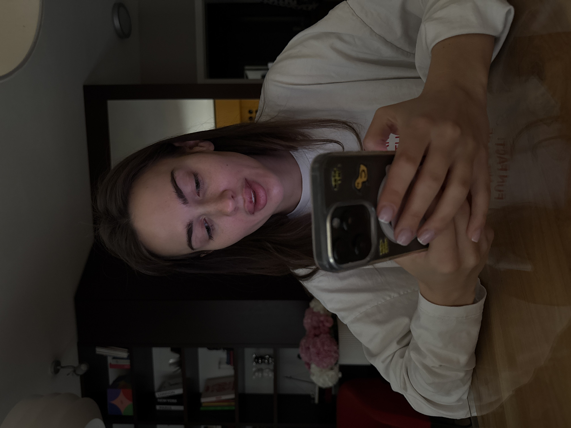 A young woman holds a smartphone in her hands, her face appearing in the frame as she seems to be taking a selfie or recording a video. She is wearing a light-colored, long-sleeved shirt. The background suggests an interior setting, possibly an apartment, with a bookshelf filled with books and decorative objects visible. The presence of what appears to be a radiator and curtains indicates a domestic environment. While there is no direct indication of Warsaw, Poland, from the visual details alone, the setting could reasonably be a modern apartment in an urban area, matching the characteristics of a city like Warsaw.