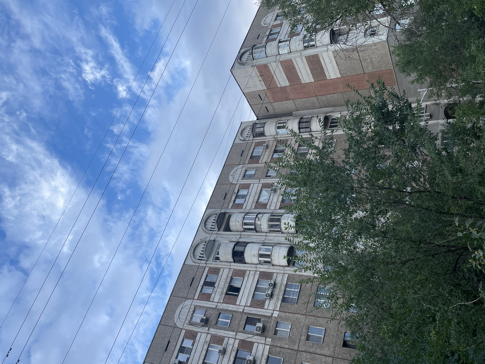 This is a low-angle, eye-level, medium shot of a multi-story apartment building in Almaty, Kazakhstan, viewed from a street-level perspective. The building, constructed from concrete and concrete panels, exhibits a grey and pink color scheme with arched balconies and numerous windows. Several air conditioning units are visible mounted on the exterior. The sky above is a mixture of blue and white clouds, with a prominent set of power lines running diagonally across the frame. Dense green foliage from trees partially obscures the lower portion of the building, suggesting an urban park or residential area with established landscaping. The overall impression is that of a typical residential complex in a city setting.