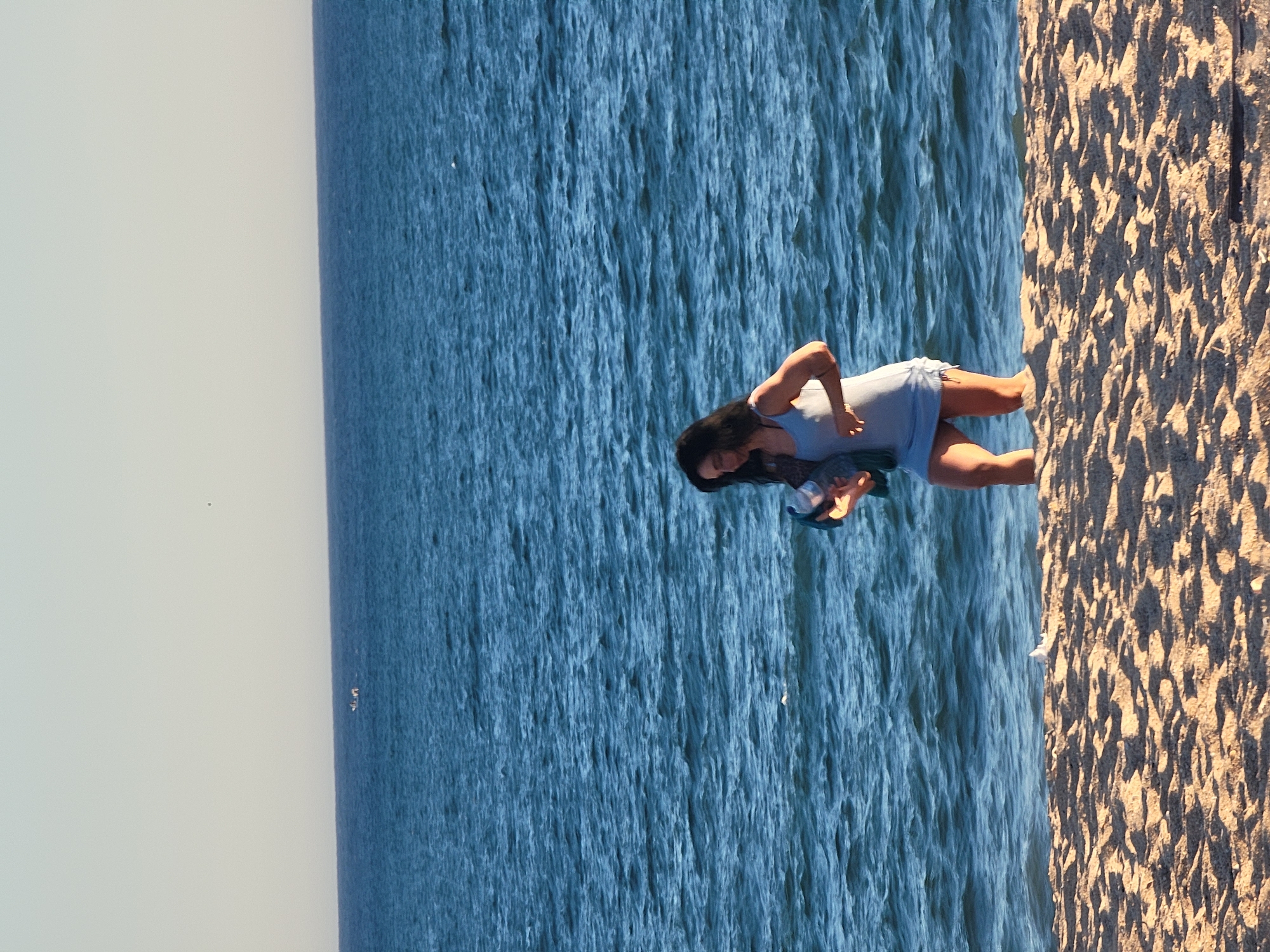 In Quarteira, Portugal, a woman with dark hair is standing on a sandy beach. She is wearing a light blue dress and is holding a dark blue bag and a bottle. The ocean is behind her, with gentle waves. In the distance, on the water, a small boat with people can be seen. The sky is clear and a light shade of yellow and white.