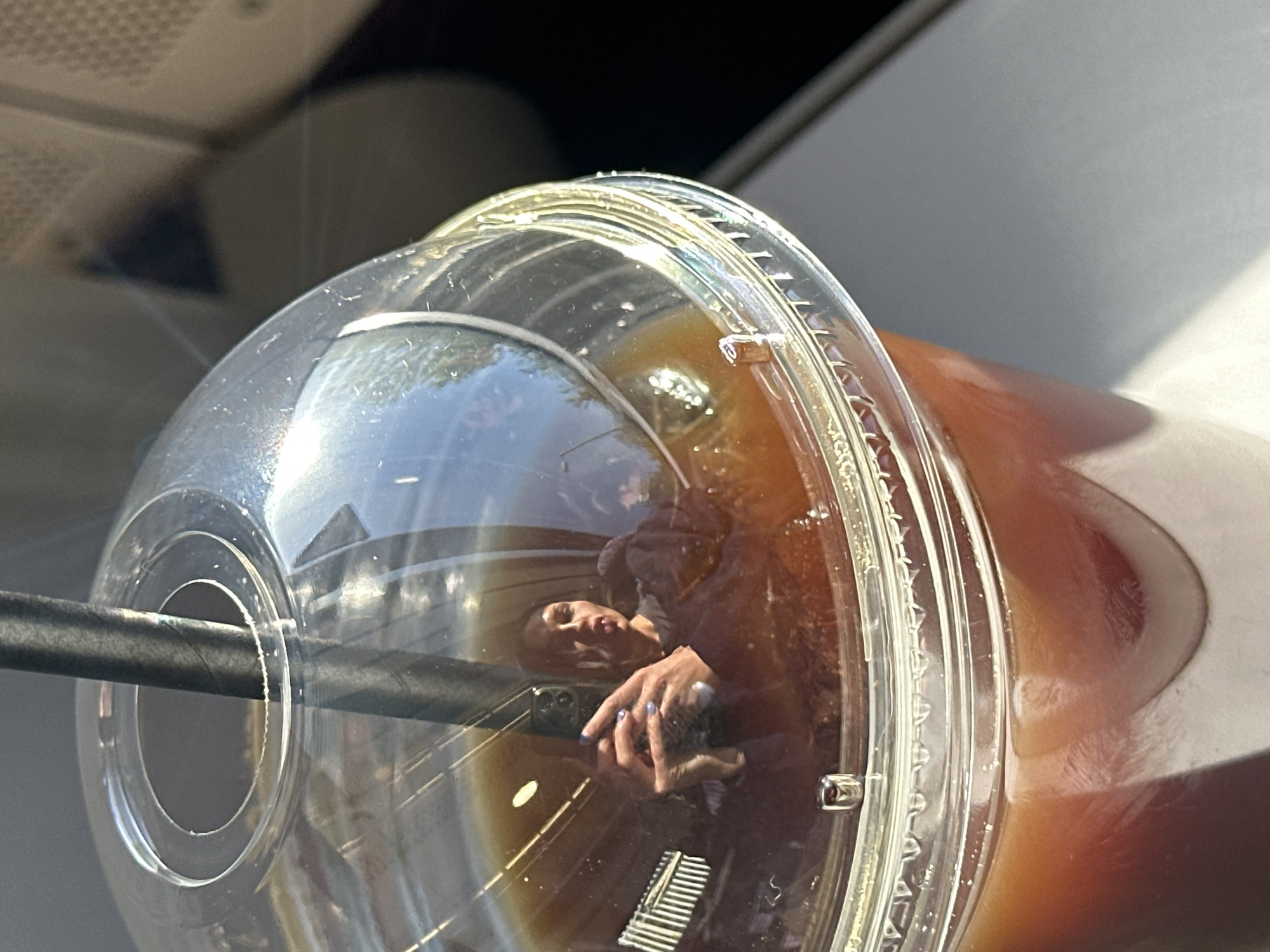The image shows a close-up of a plastic cup filled with a dark liquid, possibly iced coffee. A black straw is inserted through the lid. The lid is clear and reflects a person holding a smartphone. The background is out of focus, but suggests an indoor setting, potentially a vehicle. The image provides insufficient evidence to determine the location with certainty. Therefore, I cannot confirm it to be in Tunbridge Wells, United Kingdom.