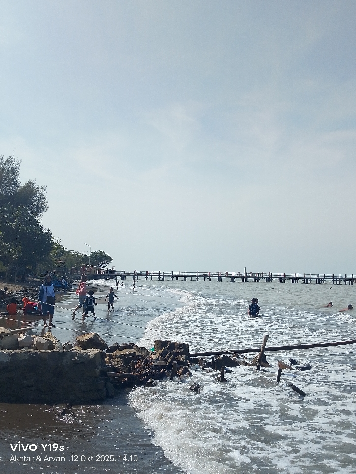 This image depicts a coastal scene, presumably in Limbangan, Indonesia, captured on October 12, 2025, at 14:10, by a vivo Y19s device. The scene is illuminated by a bright, hazy sky that occupies the upper two-thirds of the frame.
The foreground shows the ocean with moderate waves breaking into white foam near the shore. The water appears dark and murky, containing numerous pieces of debris, including logs and branches. The immediate shoreline consists of rugged rock and concrete fragments, mixed with accumulated debris.
In the midground, to the left, a sandy-muddy beach area transitions from the water. Sparse vegetation and a line of trees are visible along the landward edge of the beach. Several individuals are present in this area: one adult wearing a light blue top and dark pants stands in the shallow water, facing right. Two children are actively running through the surf; one wears a light pink top, and the other is in darker clothing. Other indistinct figures are visible on the wet sand further left. A light pole stands on the beach before the pier structure begins.
Further out in the midground water, one individual in dark clothing is partially submerged, facing away from the camera. Two additional individuals are visible further offshore, appearing to swim or wade.
A long, wooden pier extends from the midground shoreline into the distant ocean. The pier features railings along its length and is supported by numerous vertical posts. Several small, indistinct figures are discernible on the pier. The horizon is visible where the hazy sky meets the ocean surface in the far background.