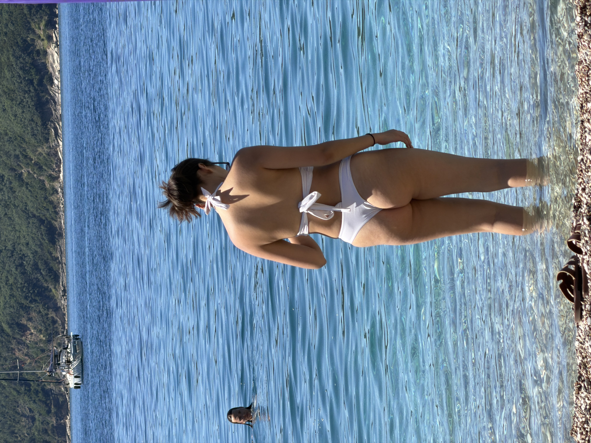 A medium, vertical shot features a woman in a white bikini standing in clear, blue water near a pebble beach. The beach is lined with dark pebbles on the right. Her backside is facing the camera. She has dark hair that is pulled back. Another person is visible in the water some distance away and to the left of the woman. A steep, green, tree-covered coastline runs along the left side of the image. A boat and other marina infrastructure can be seen near the coastline in the distance. The sky is a light blue at the top of the image. The location is identified as Bečići, Montenegro.