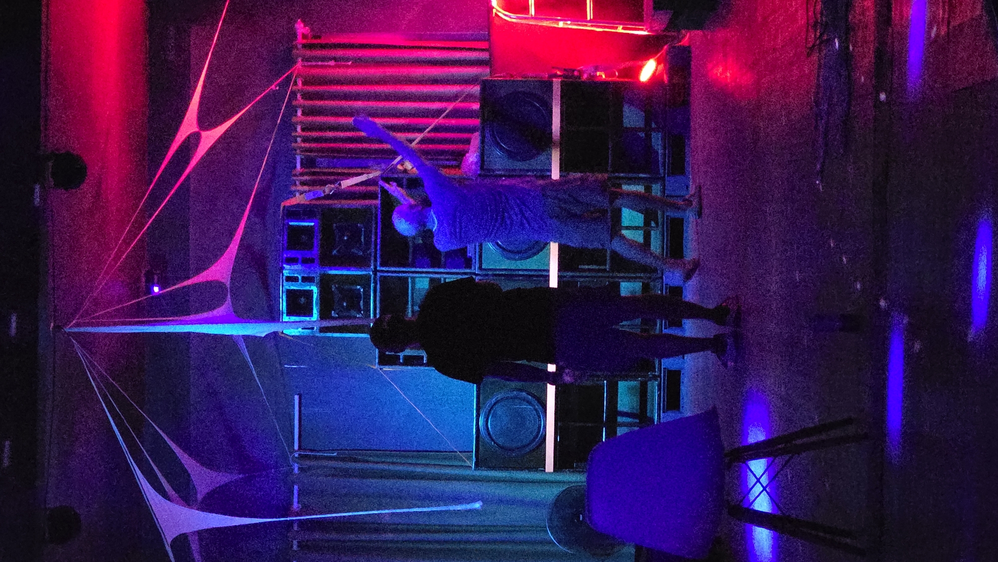 The image depicts an indoor scene in Nha Trang, Vietnam, characterized by vibrant neon lighting, primarily in shades of red, blue, and purple. A stack of speakers is prominently featured, with two individuals positioned in front of them. One person is in a light-colored shirt, raising an arm, while the other is in a dark shirt. A chair sits on the right side of the frame, also illuminated in blue light. A web-like structure is on the left.
