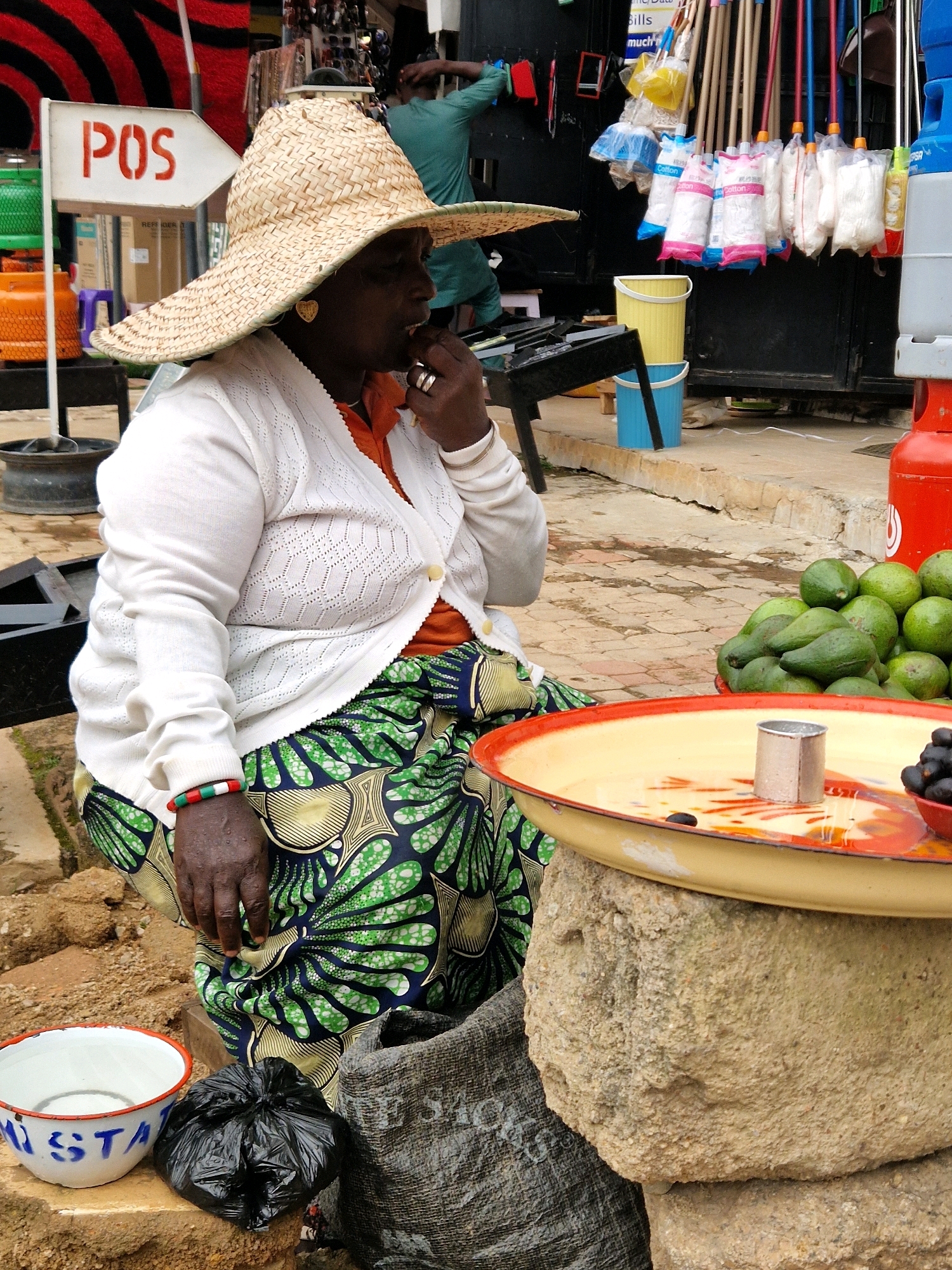 The image depicts a street scene in Jos, Nigeria. A woman, likely a vendor, is seated wearing a wide-brimmed straw hat, a white knitted cardigan over an orange shirt, and a vibrant green and blue patterned skirt. She has her hand to her mouth, seemingly eating or tasting something. In front of her, a large, shallow tray with an orange rim holds a small metal cup and some dark berries. To the left, a white enamel bowl with blue lettering is visible, along with a black plastic bag. Behind her, a pile of green avocados is displayed. The background reveals stalls with various items for sale, including what appear to be cleaning supplies like mops and buckets, and gas cylinders. A sign with "POS" in red letters is also visible. The ground is paved with interlocking bricks. The overall atmosphere suggests a busy market environment.
